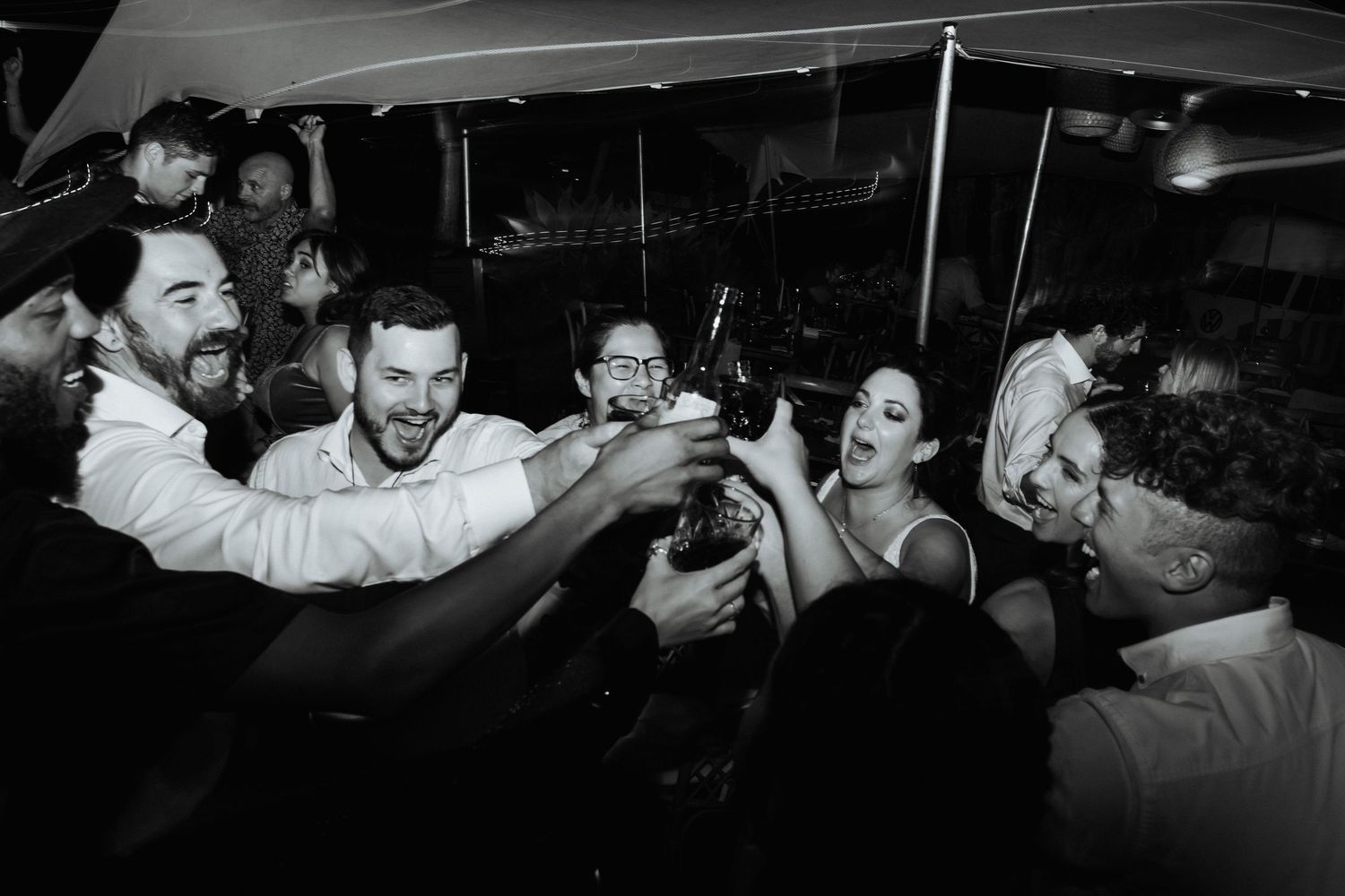 Black and white image of a drunk bride and groom smiling and cheering with their guests in the late evening