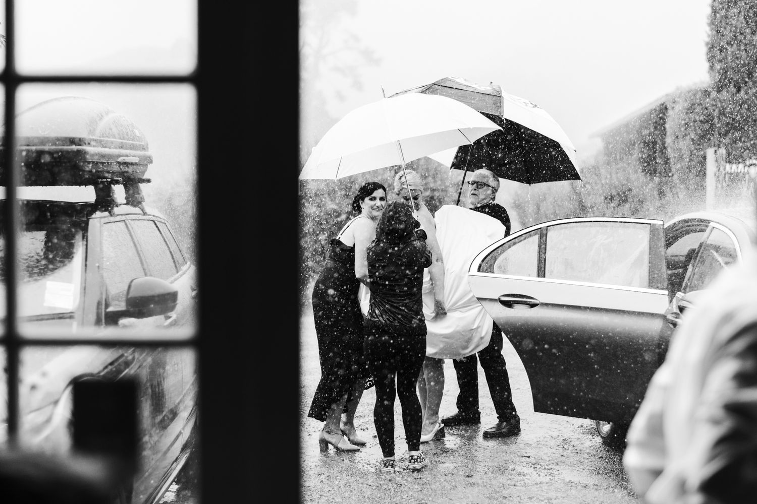 The bride gets out of the car in torrential rain being helped by three people with umbrellas at Poderi Crisci