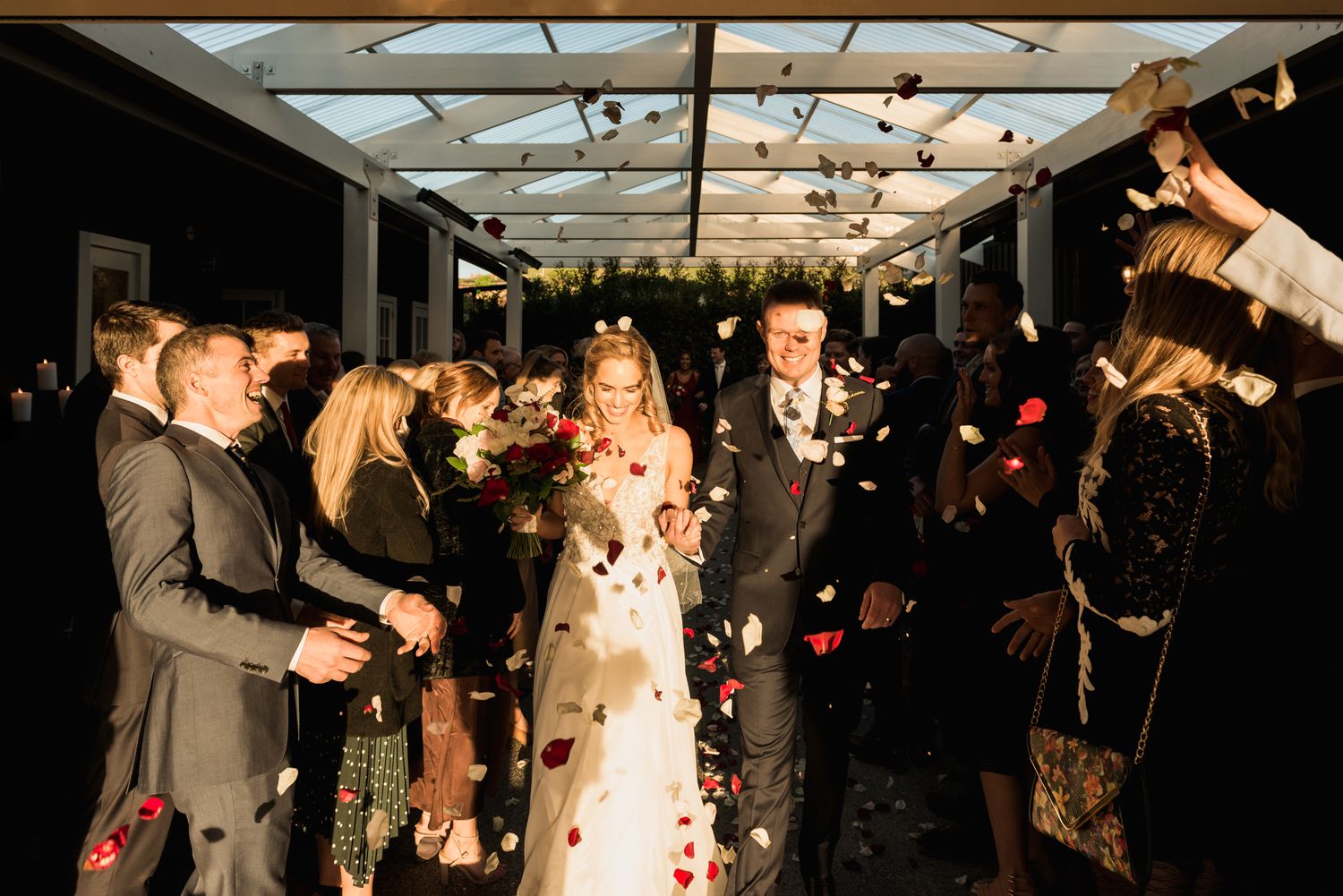 a bride and groom walk towards the camera with flower petals in white and red thrown over them, the light catches them glowing at Mudbrick