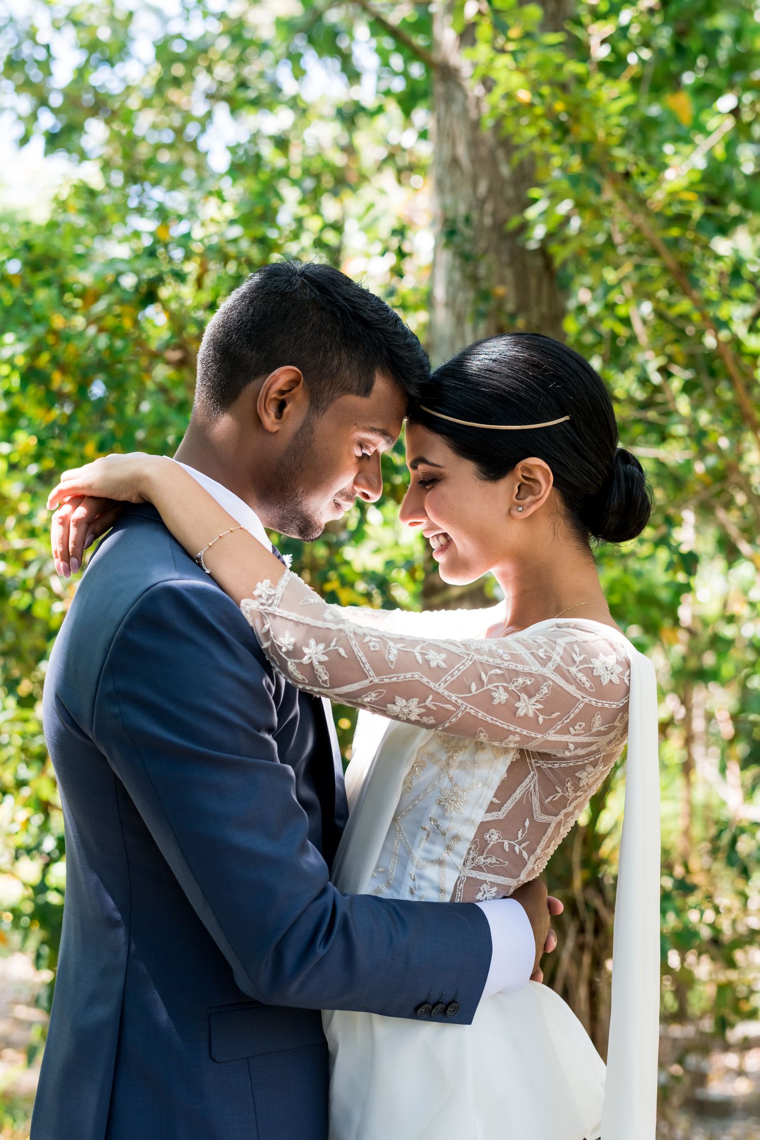 A bride and groom touch foreheads and casually drape their arms around each other under some trees in dappled light