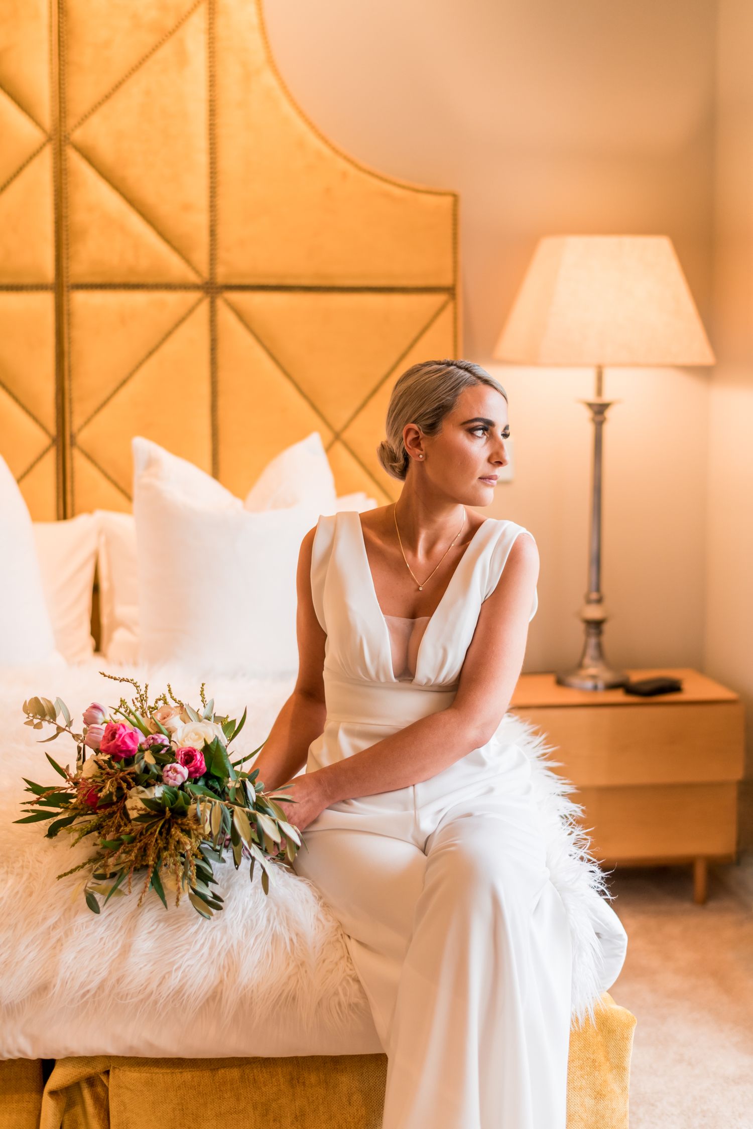 a bride sits on the edge of the bed in one of the bedrooms at Mudbrick, ornate golden velvet headboard and lamp create an image of glamour