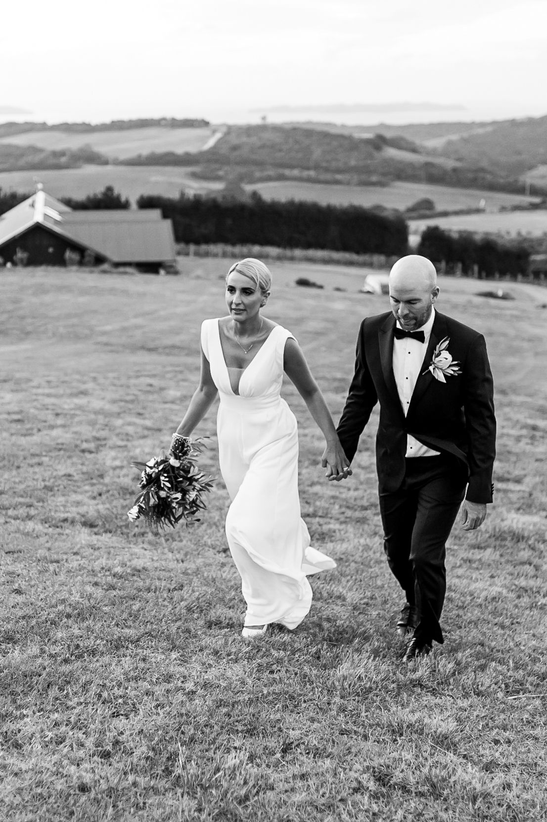 a black and white image of a bride and groom naturally walking up a hill towards the camera holding hands