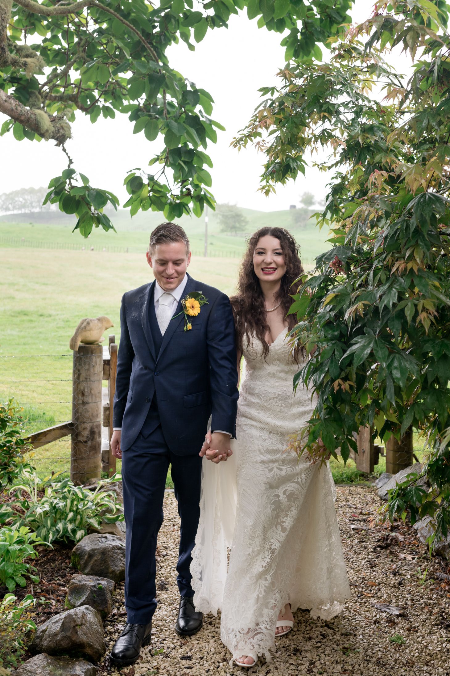a bride and groom walk towards the camera on a path through the greenery smiling and holding hands