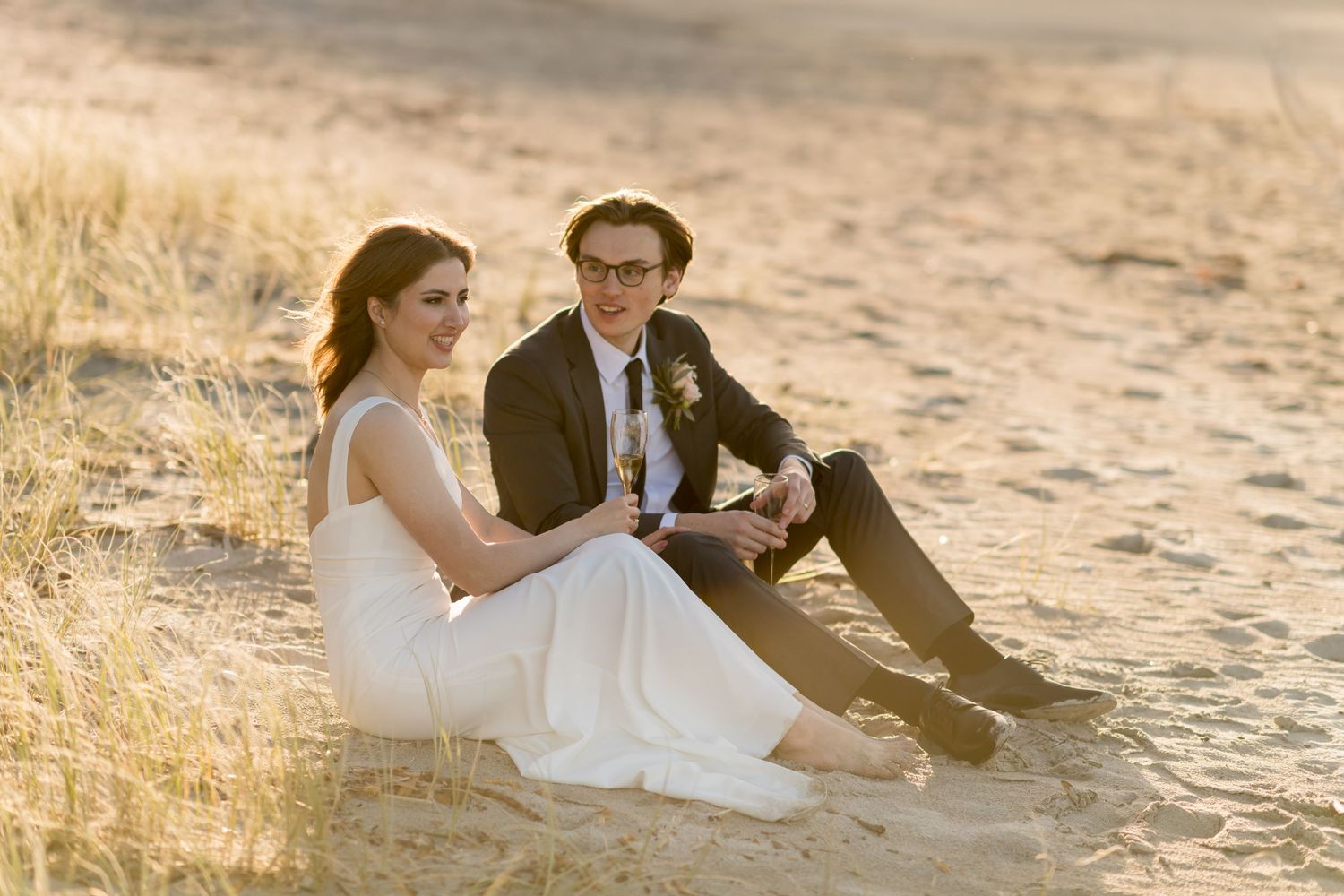 a couple newly married sit in the sand together drinking champagne in the setting sun on Onetangi beach on Waiheke Island