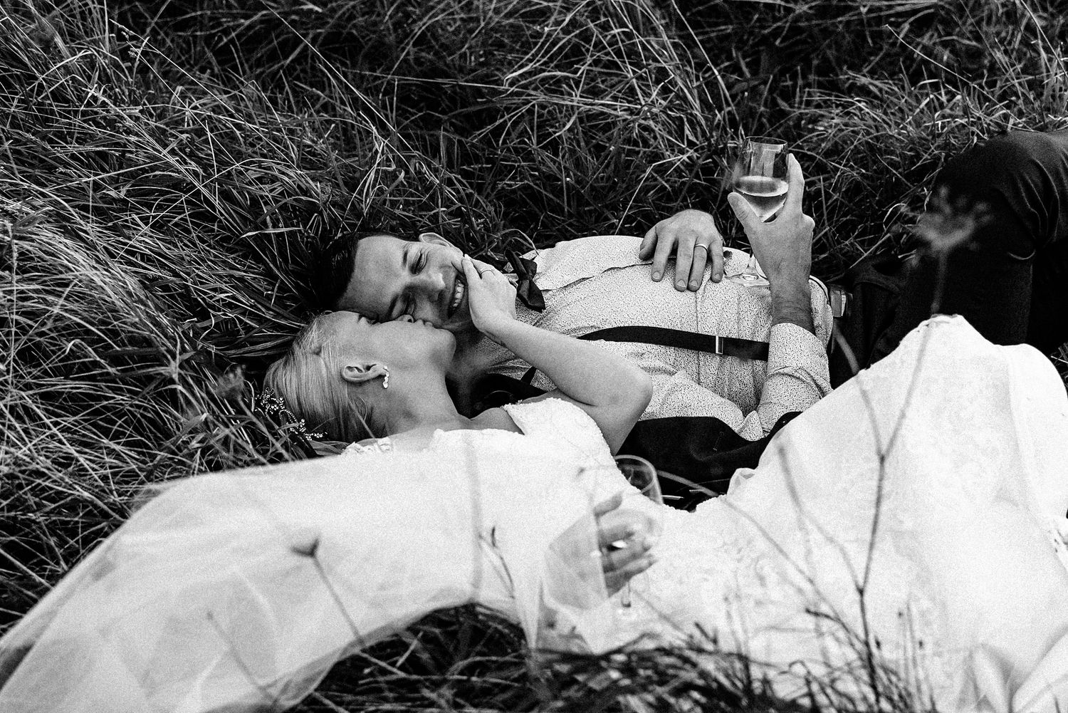 a bride and groom lay back in some long grass with a glass of wine, she gives him a pack on the cheek and he smiles