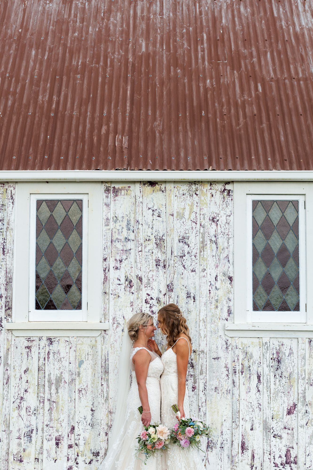 two brides embrace in front of a textured wall and glass windows from an old chapel, they mirror each other with their flowers and dresses