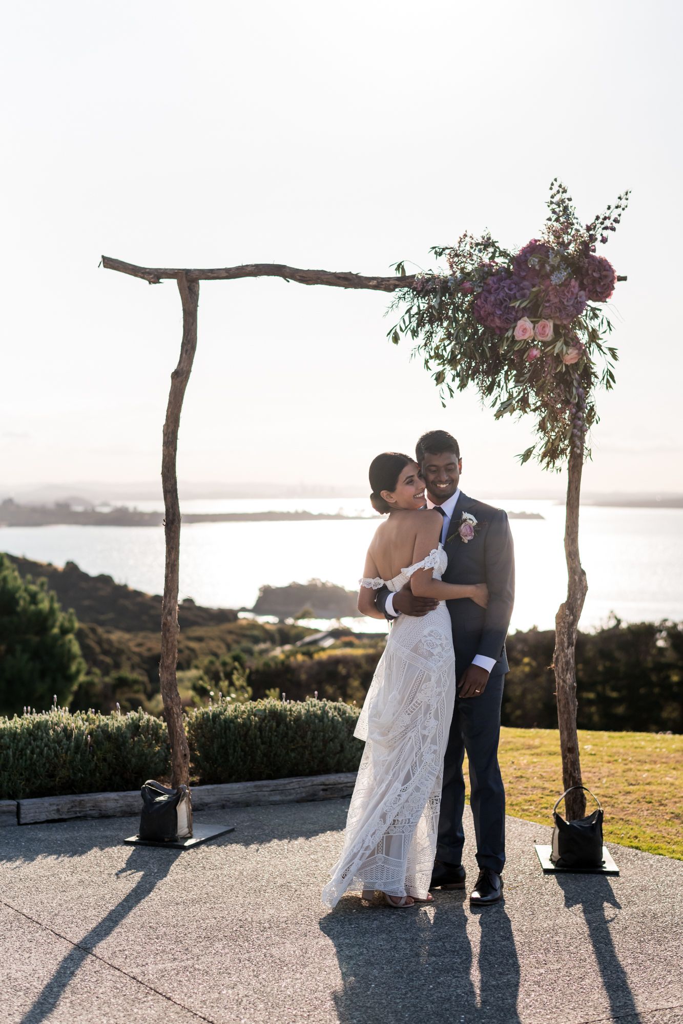 a bride and groom pose under their wedding arch with the evening sunlight, they are embraced and happy at Mudbrick vineyard