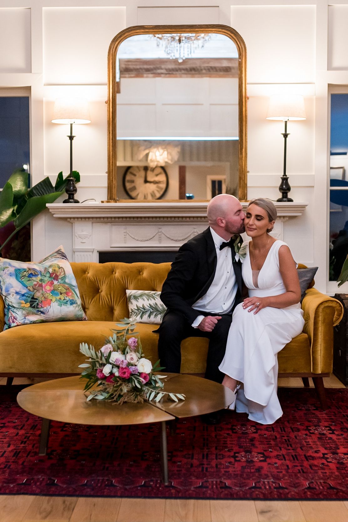 a groom pecks his brides cheek whilst they sit next to each other on a sofa in the ornate living room at Mudbrick