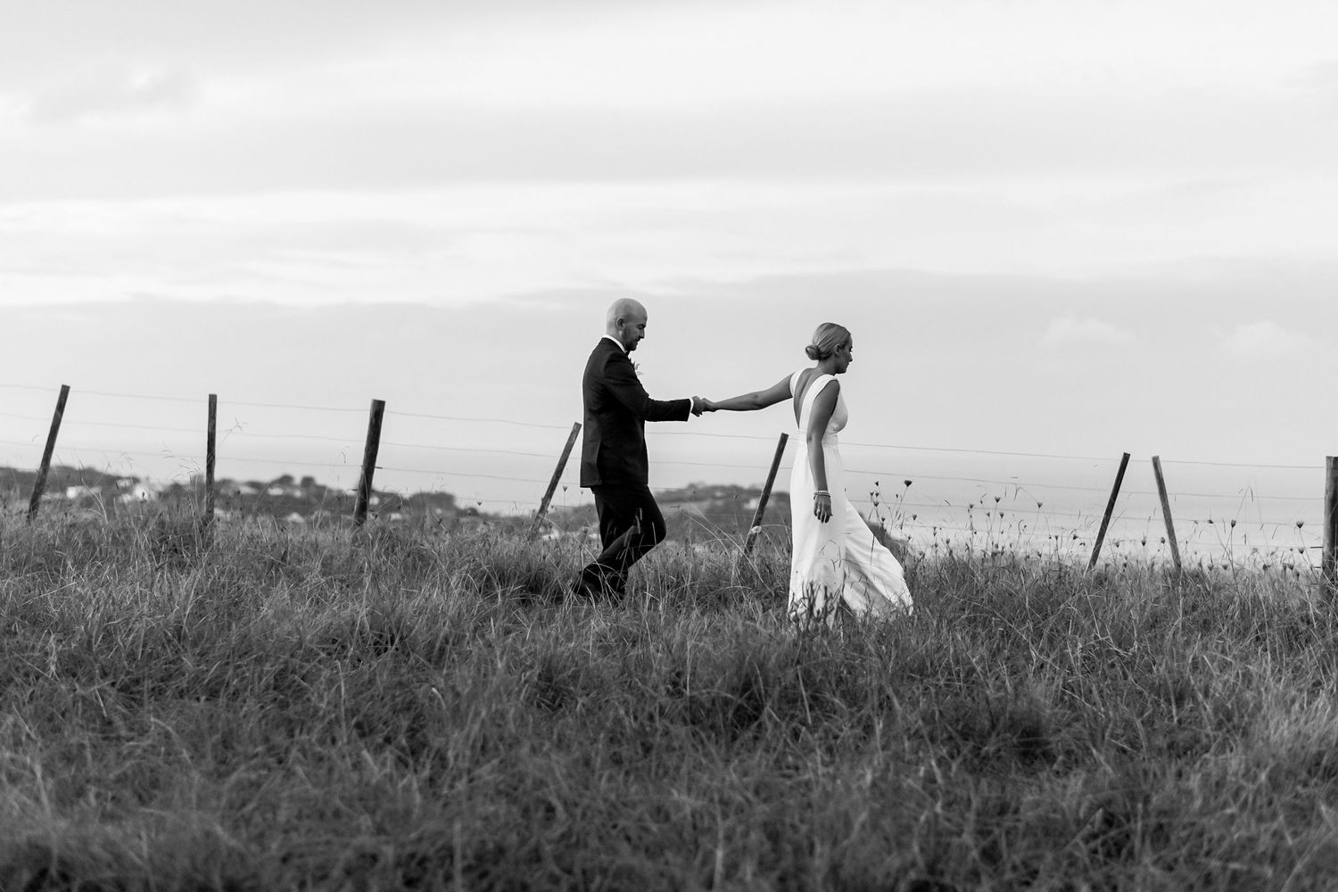 a black and white image of a bride leading the groom behind her through the long grass at the top of the Mudbrick hill