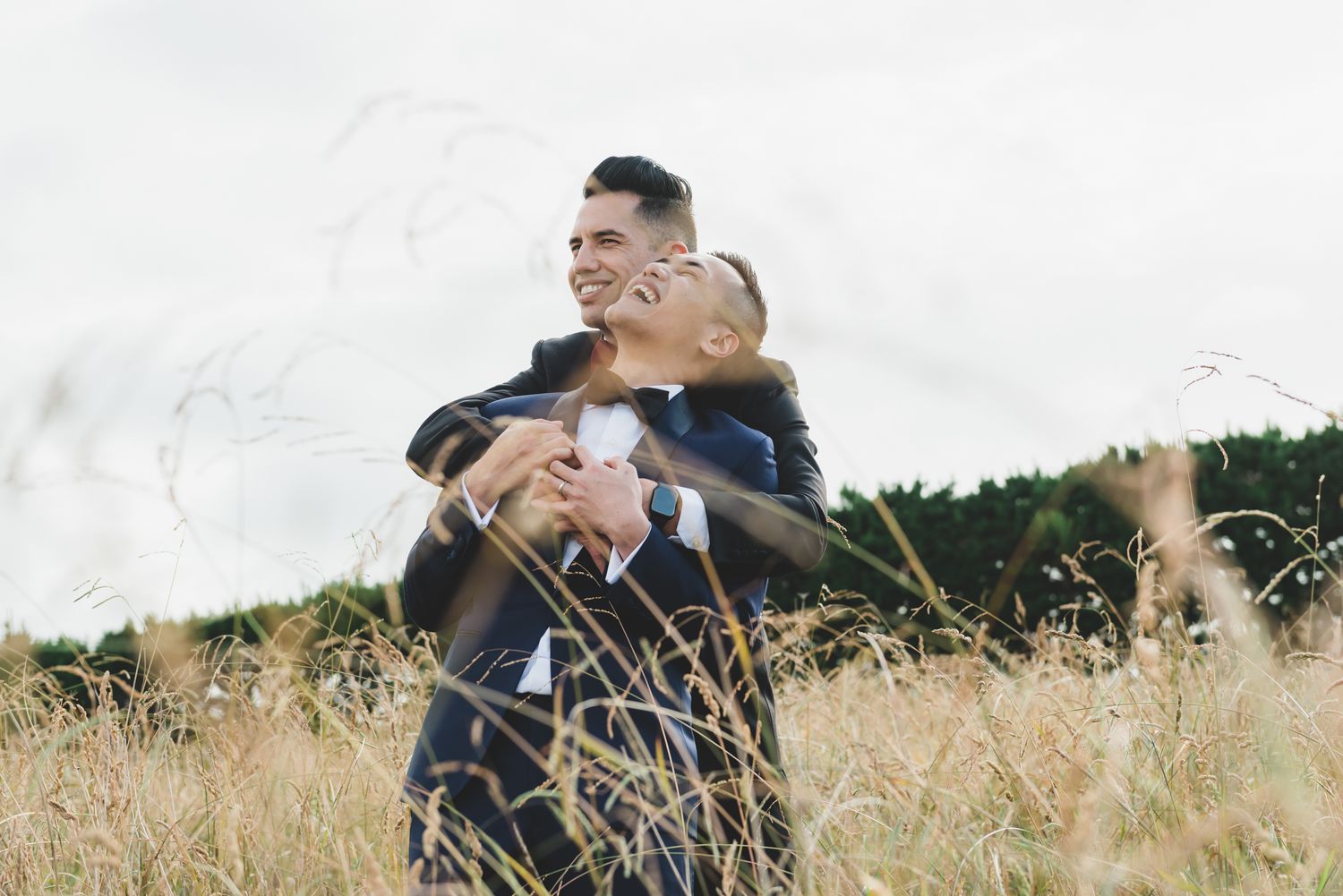 a same sex male couple on their wedding day stand in long grass the photo looks through creating patterns around them whilst they laugh