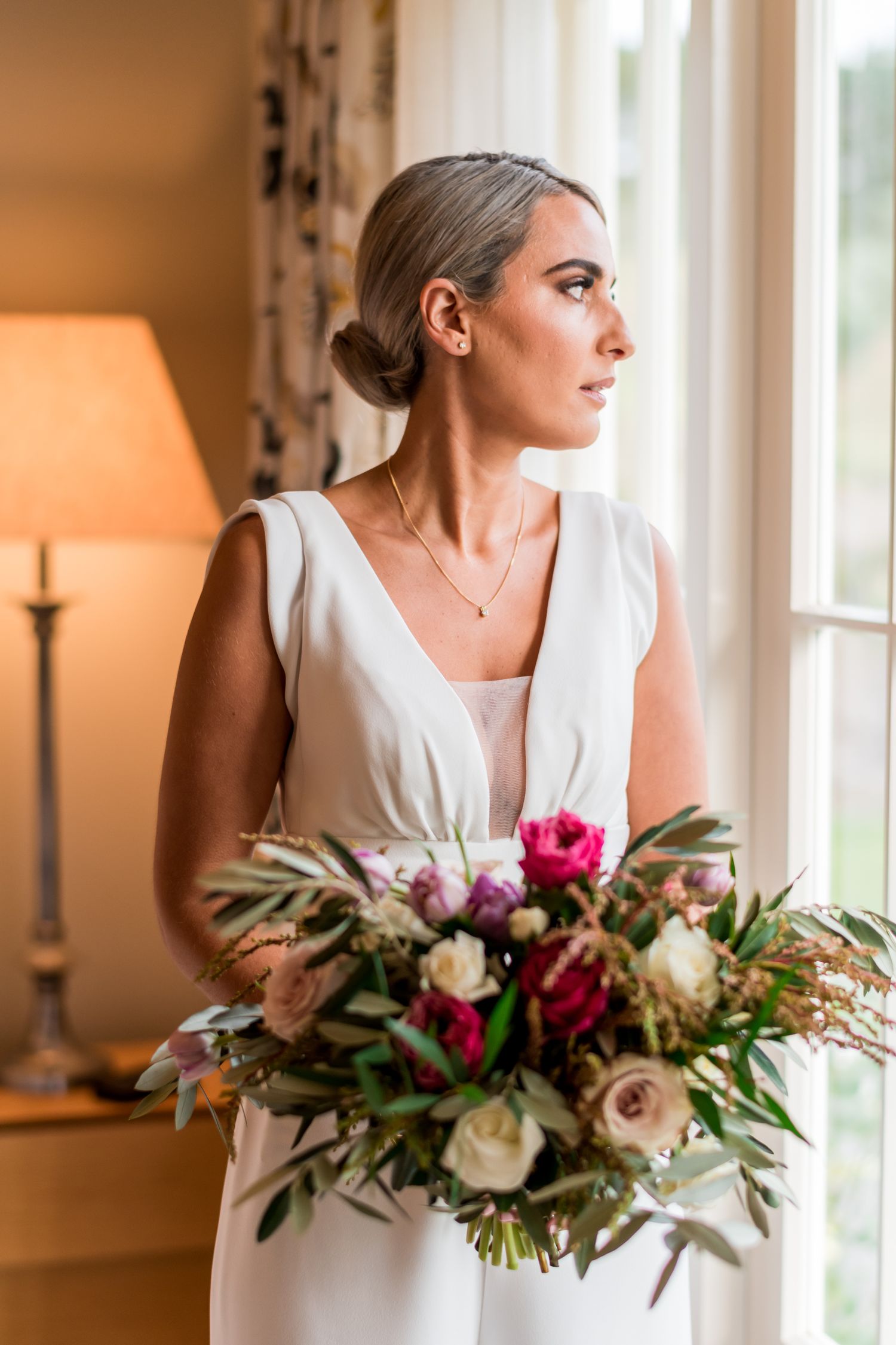 a bride who has just got ready looks out of the window holding her full bouquet of flowers thinking of her coming wedding day
