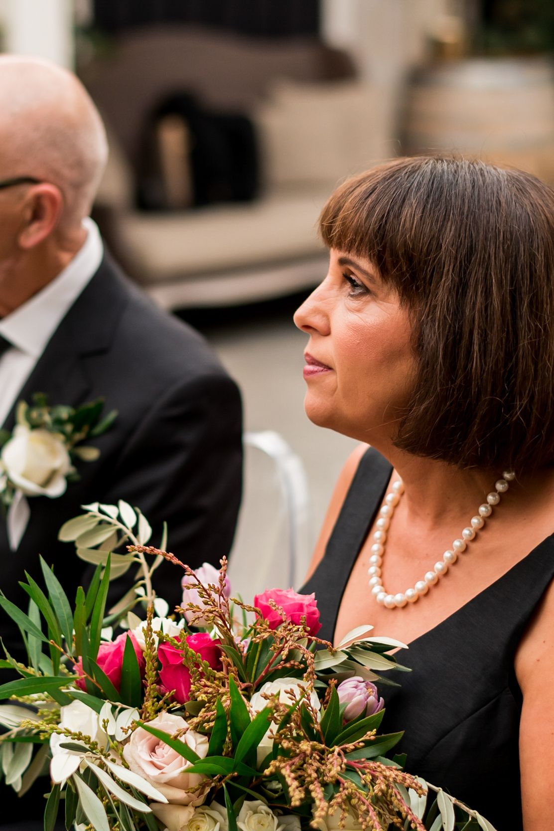 the mother of the bride sits in the wedding ceremony holding her daughters flowers whilst she gets married at Mudbrick, Waiheke.