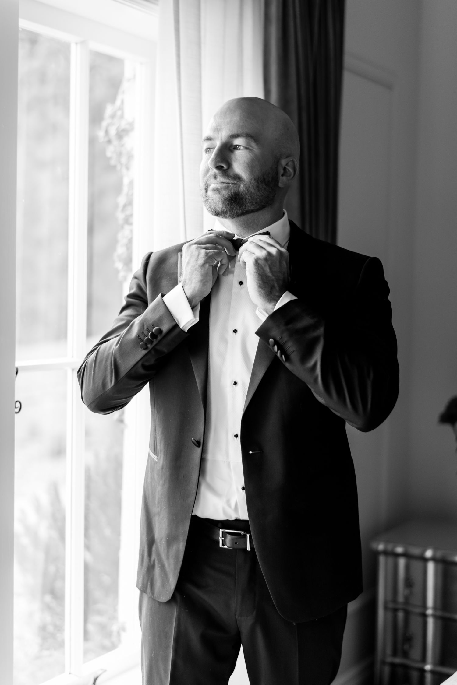 a black and white image of a groom adjusting his bow tie at the widow, he is covered in the side light light coming in