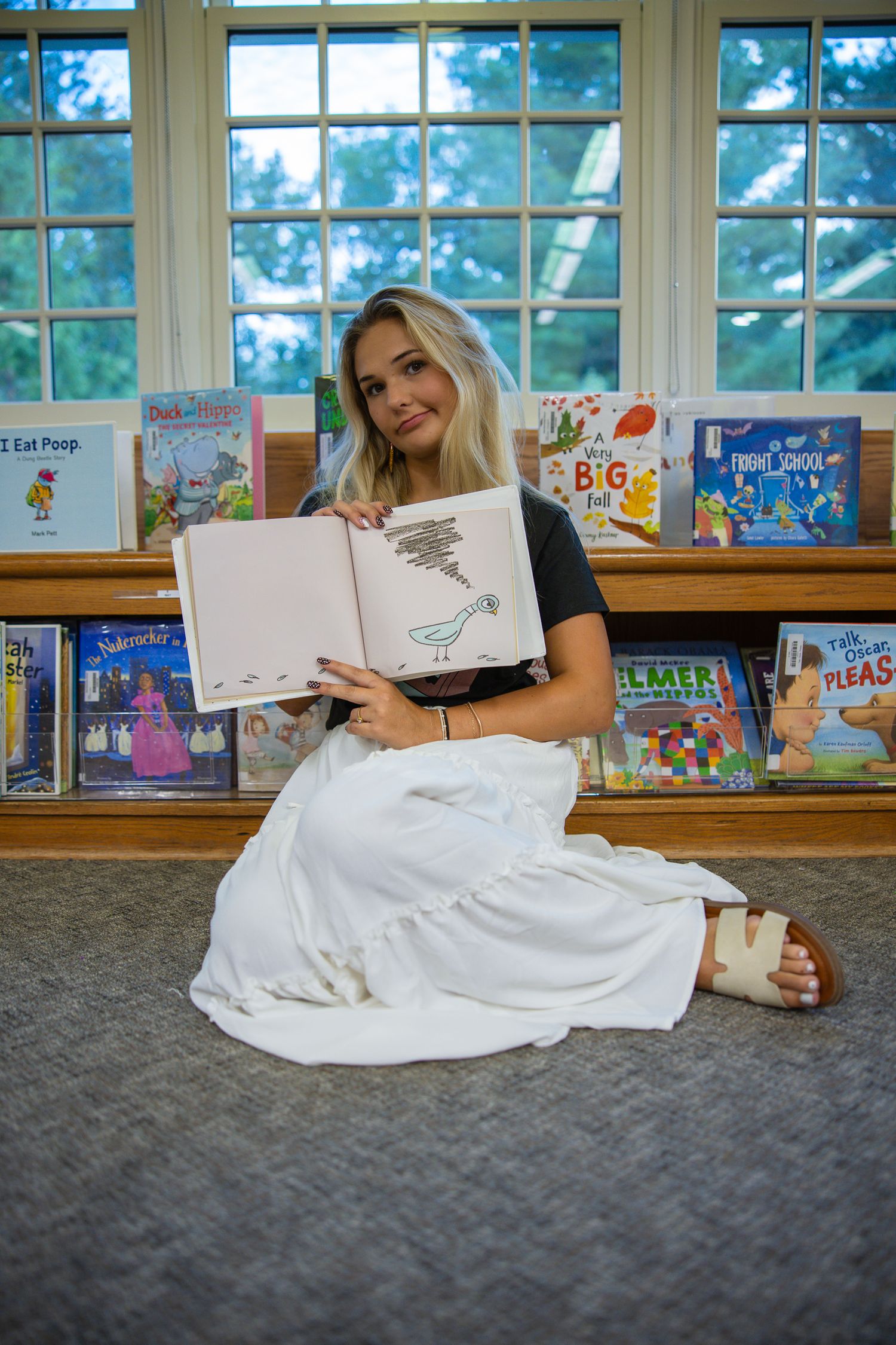 Teacher lying on library floor surrounded by children's books and school supplies.