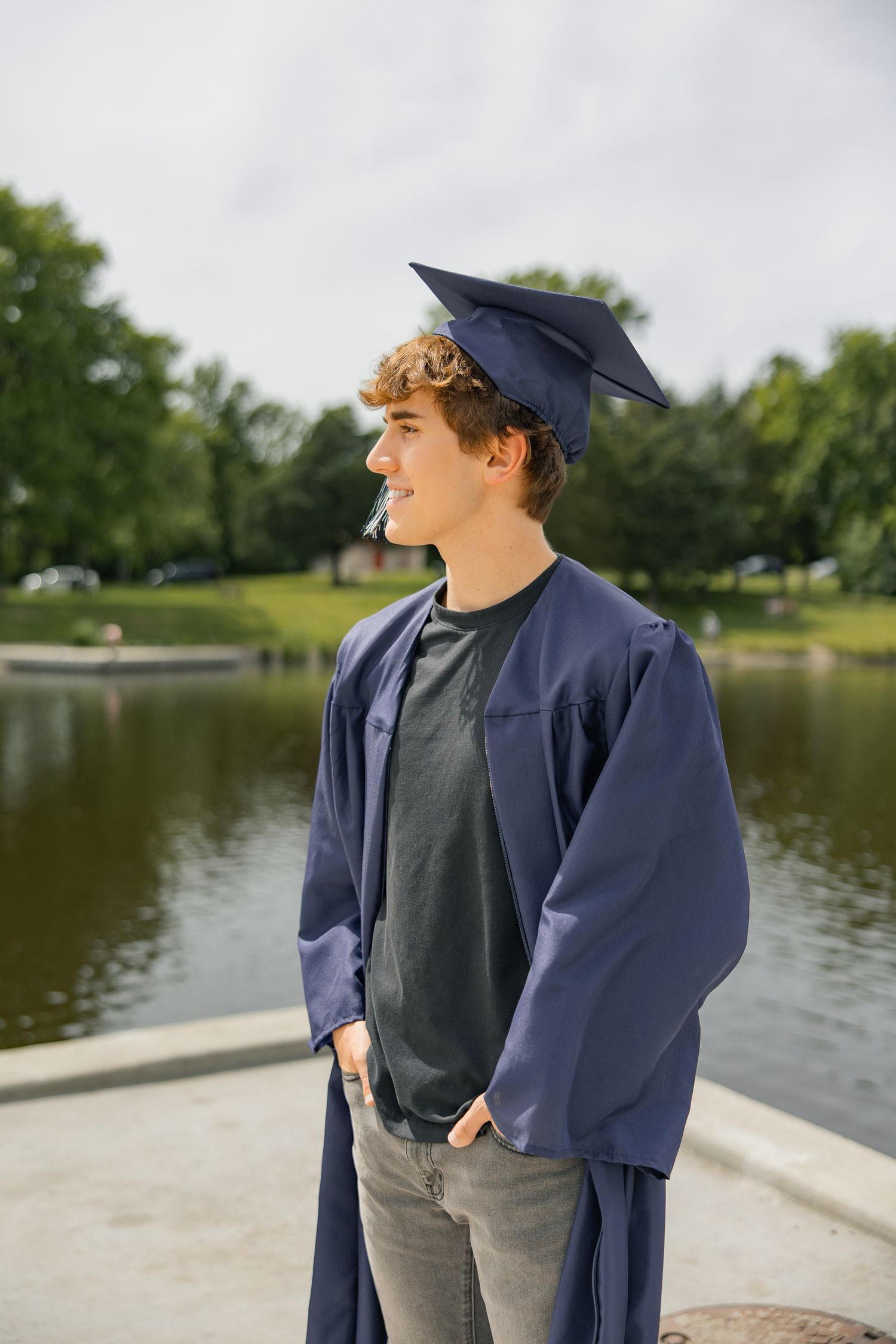 A graduate in cap and gown poses by a lake on a cloudy day.