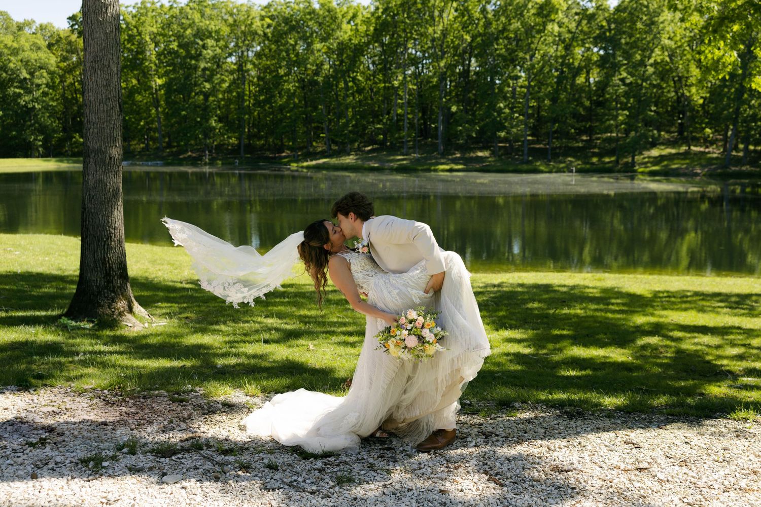 Bride and groom share a romantic dip kiss near a serene pond surrounded by lush green trees on their wedding day.