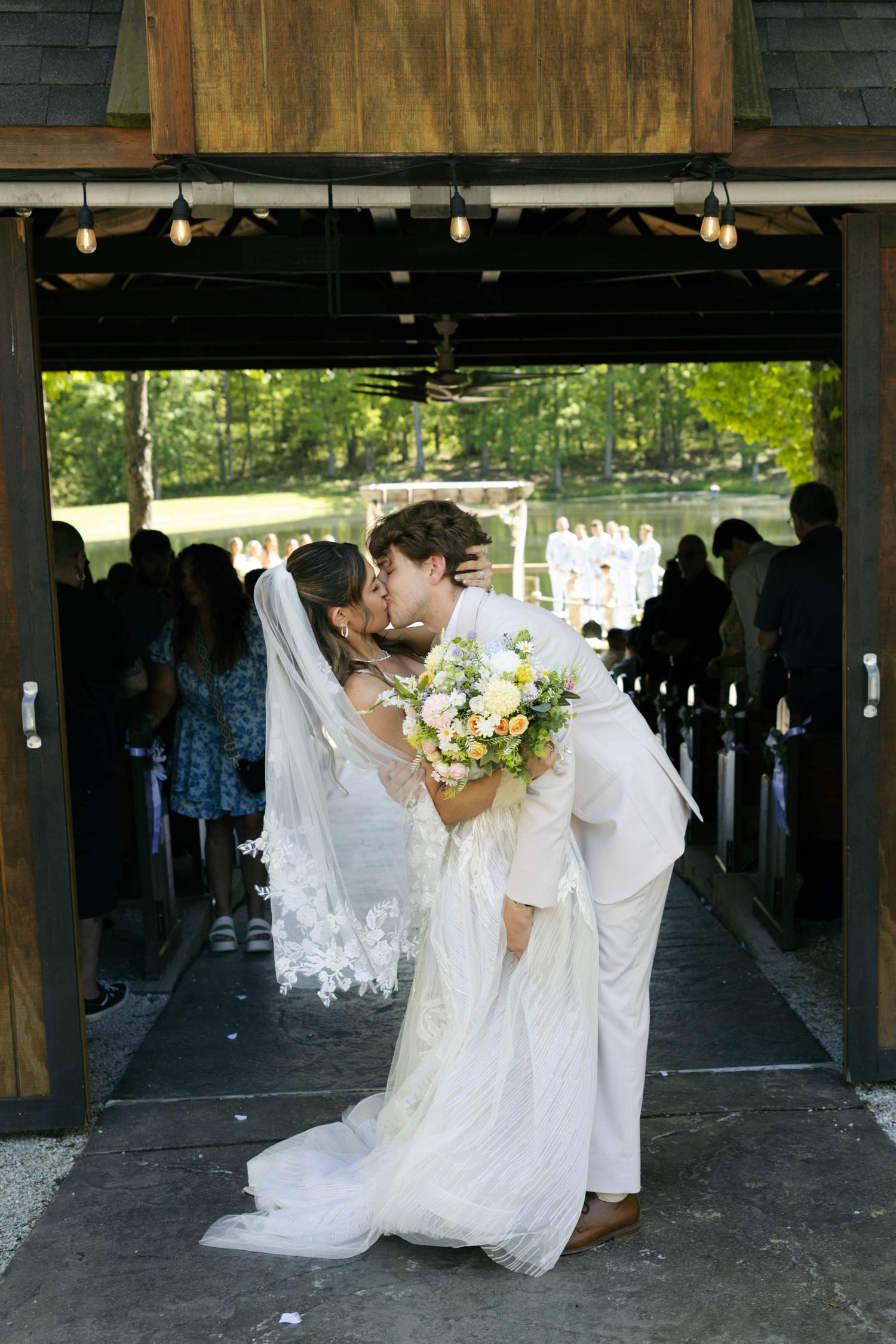 Bride and groom share first kiss at rustic outdoor wedding chapel aisle, bride holding pastel bouquet in white gown and veil.