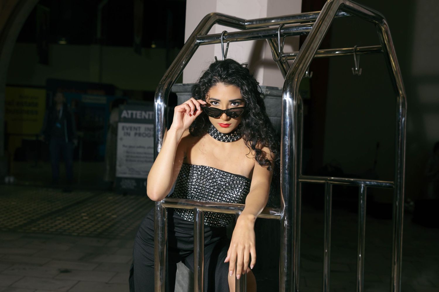 Fashionable woman in black strapless outfit and sunglasses posing inside a metal luggage cart.