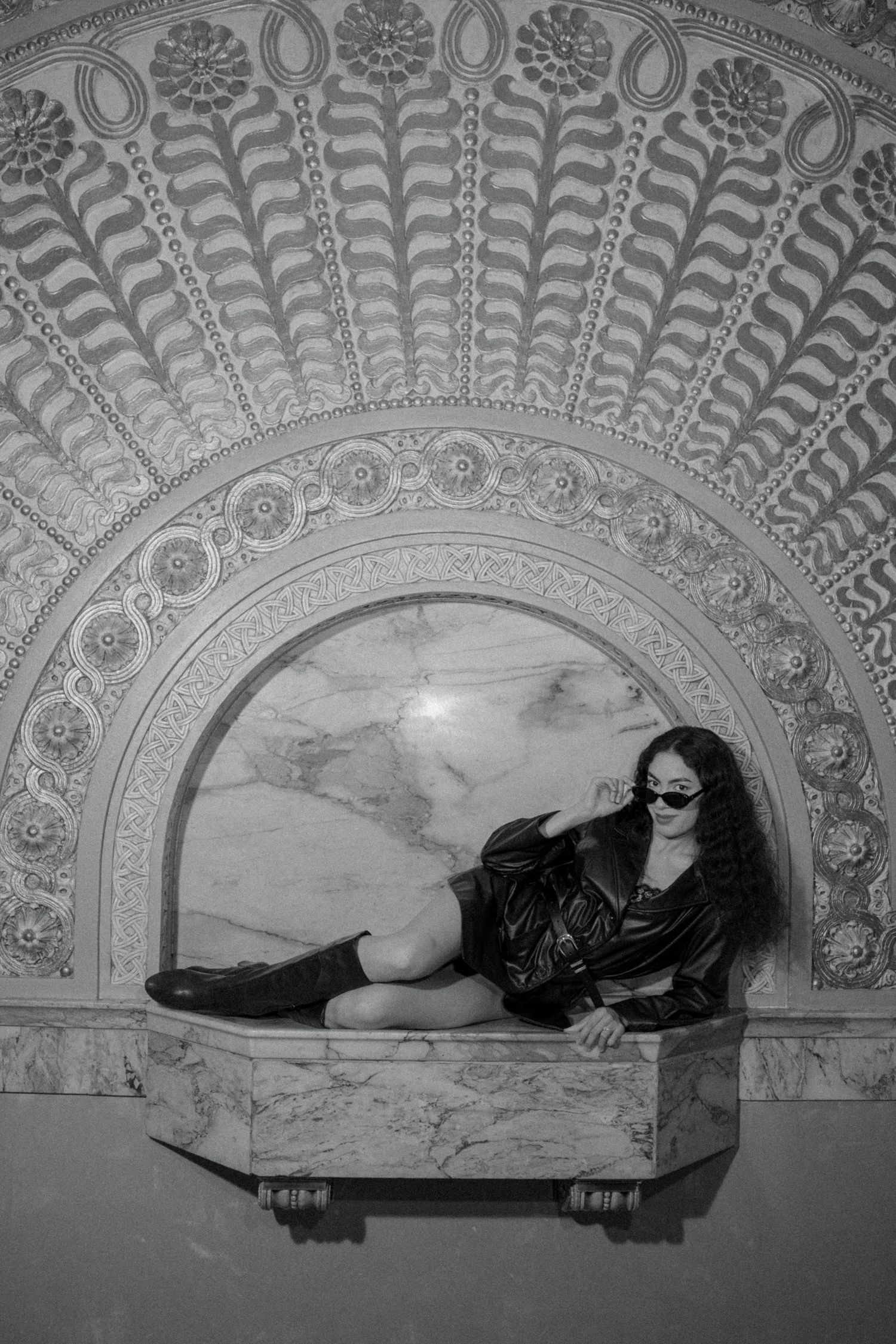 Black and white photo of a woman in sunglasses reclining on a marble ledge beneath an ornate arched relief wall.