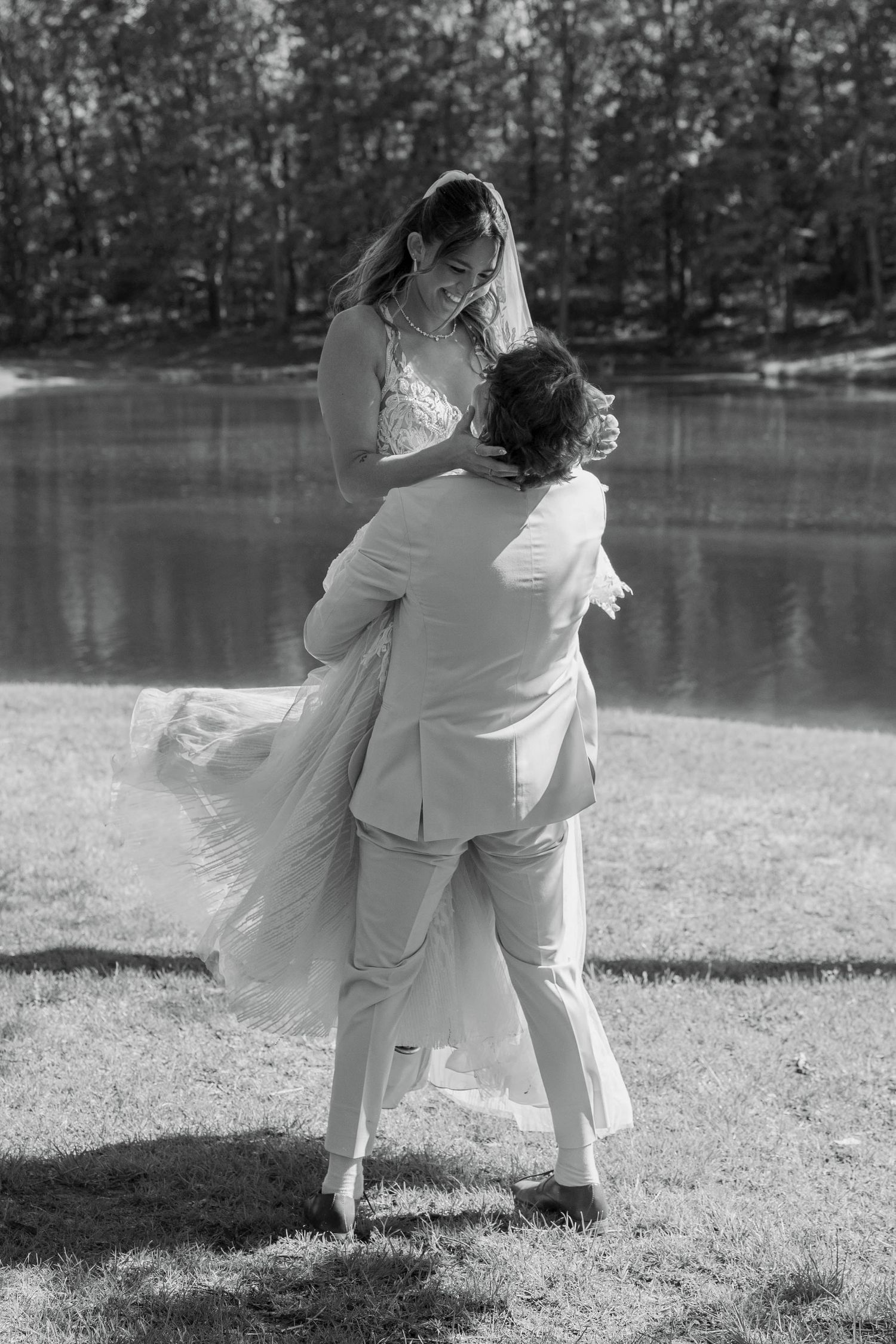 Black and white photo of groom lifting bride in wedding attire by a lake surrounded by trees.
