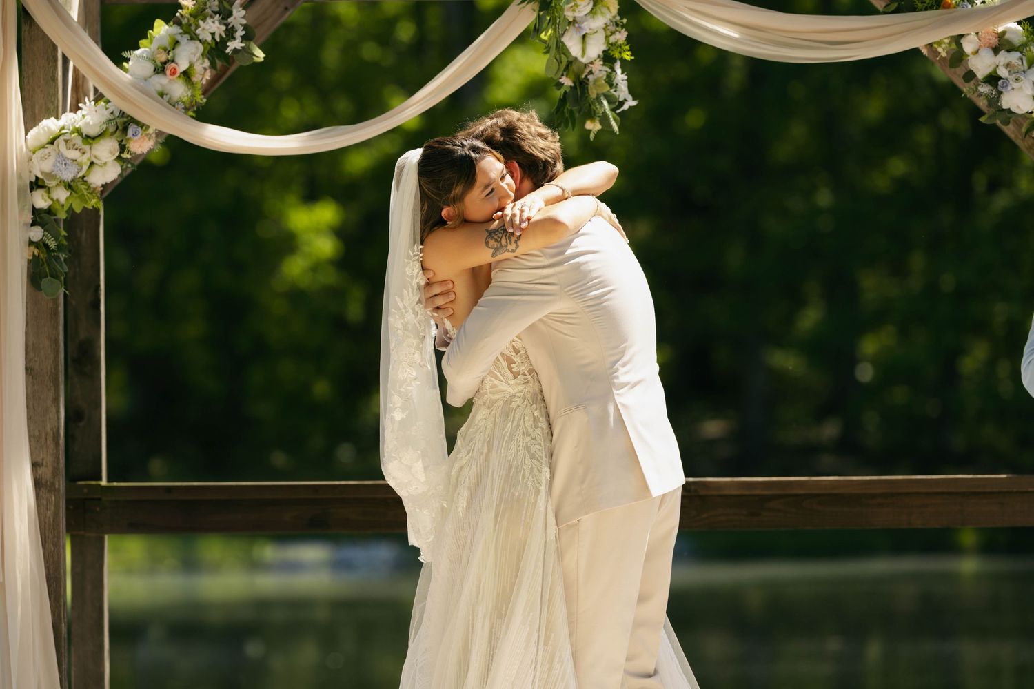 Bride and groom sharing a romantic embrace under a floral arch at an outdoor wedding ceremony by a lake.