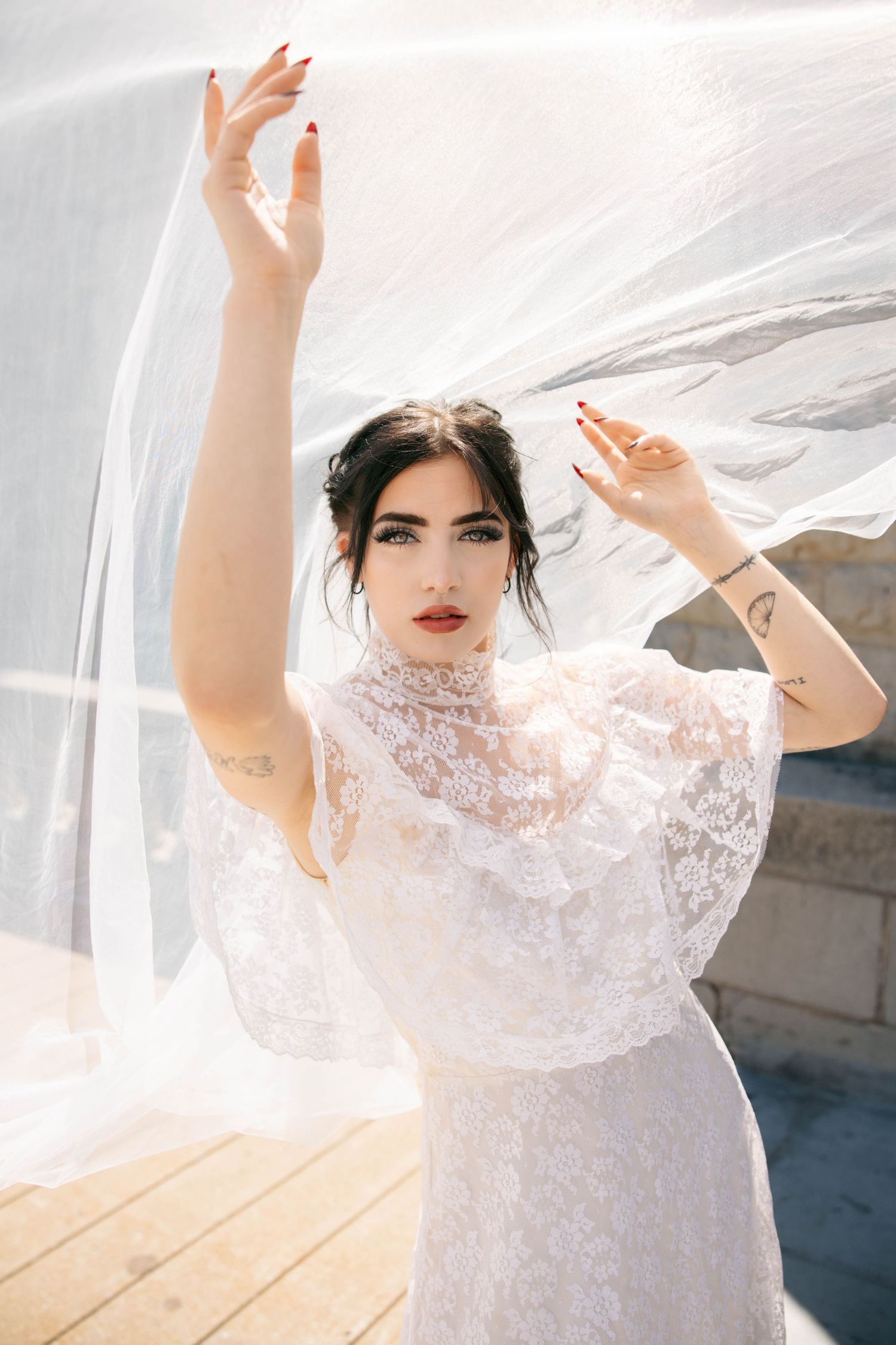 Bride in lace high-neck wedding dress lifts veil dramatically with tattooed arms raised.