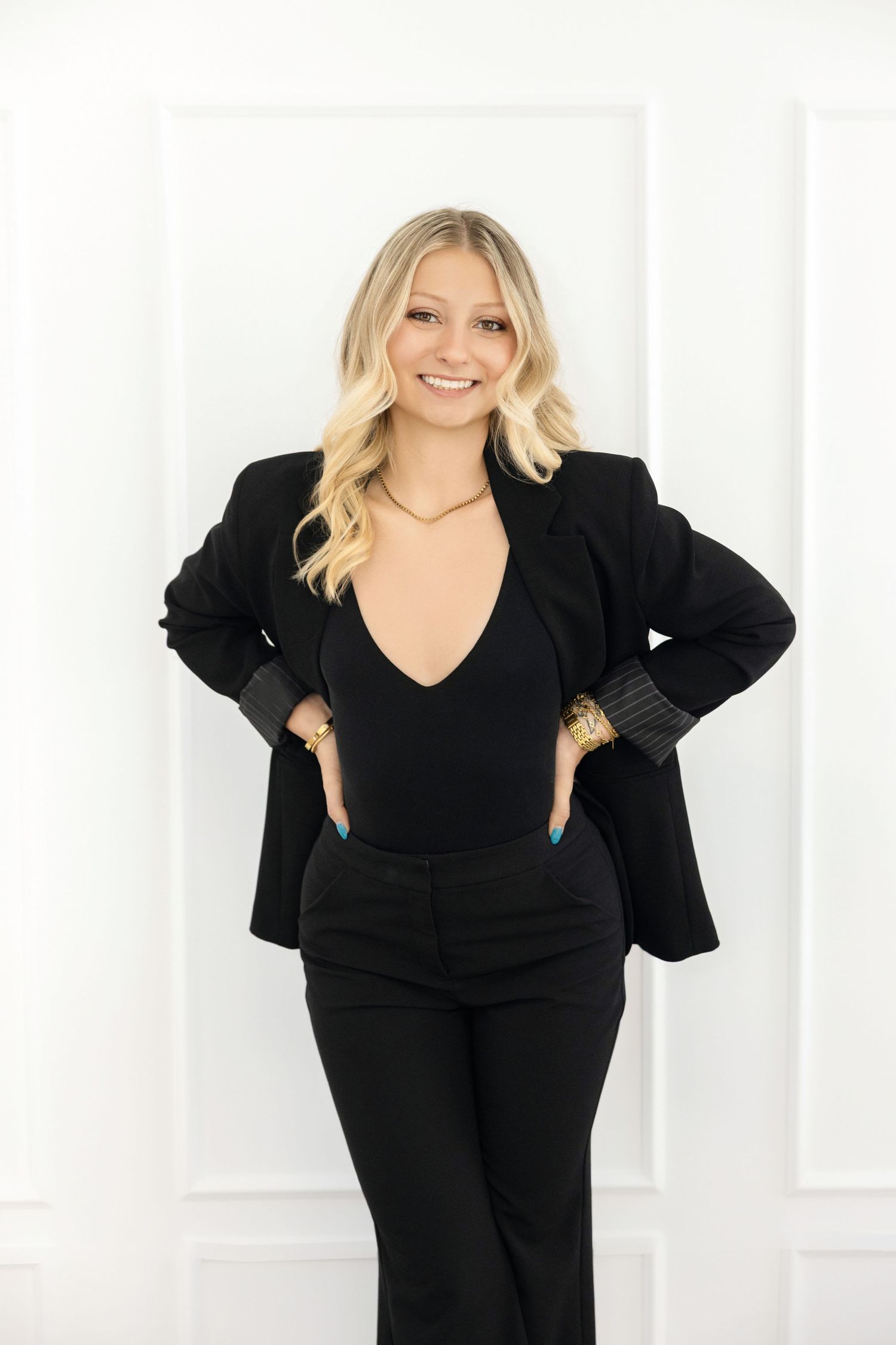 Smiling blonde woman in black blazer and bodysuit posing confidently with hands on hips against white background.