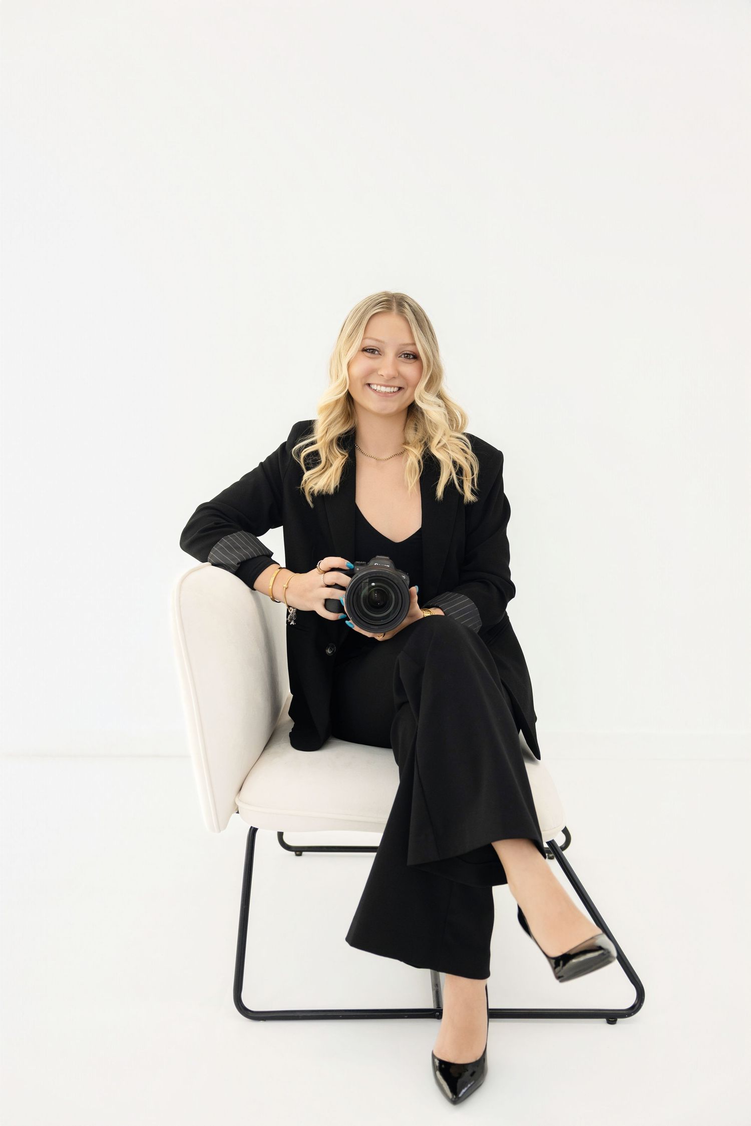 Smiling blonde woman in black suit sitting on white chair holding a professional camera against white background.