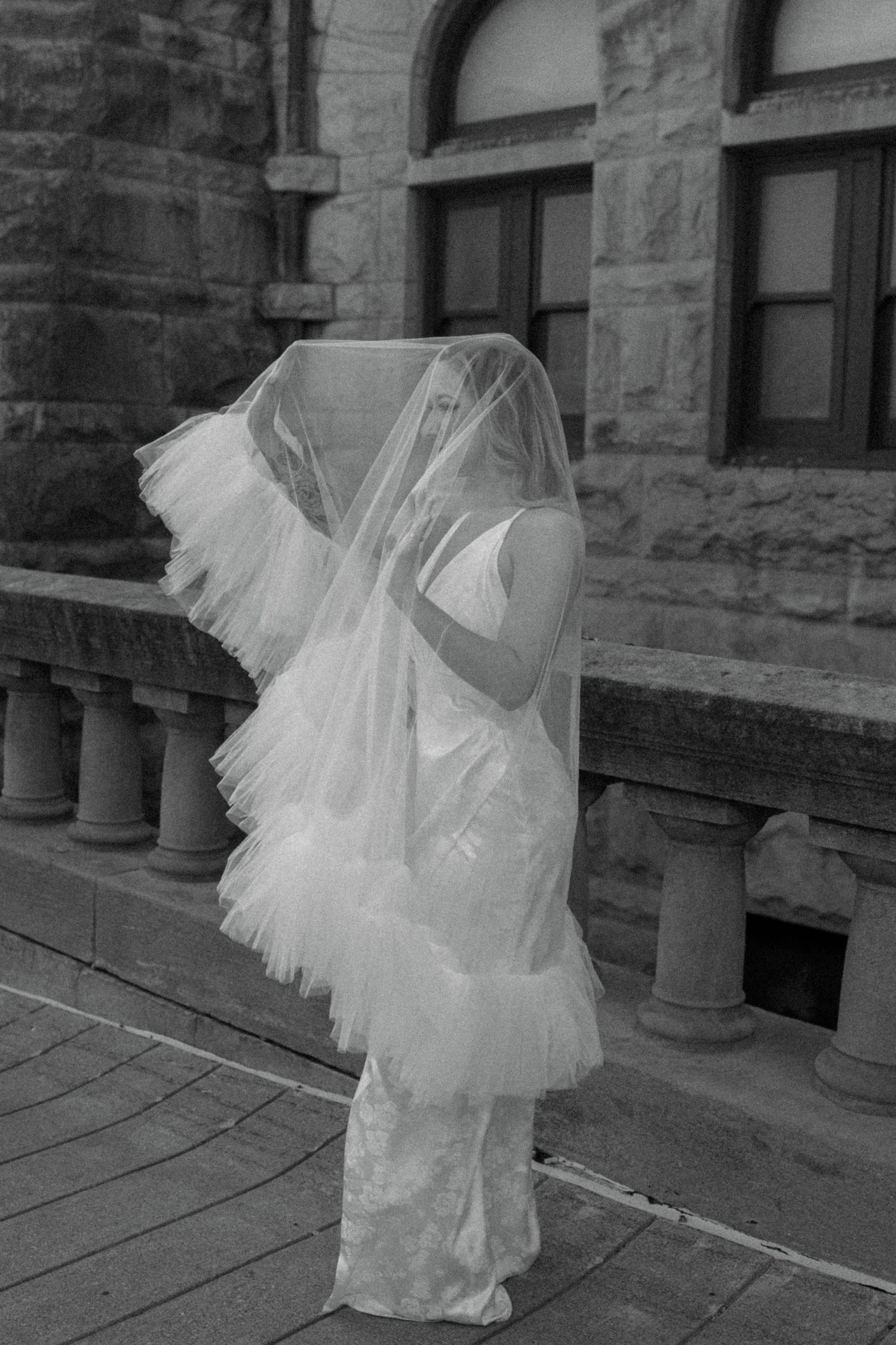 Bride in white wedding dress with flowing ruffled veil blowing over her face outdoors near stone building.