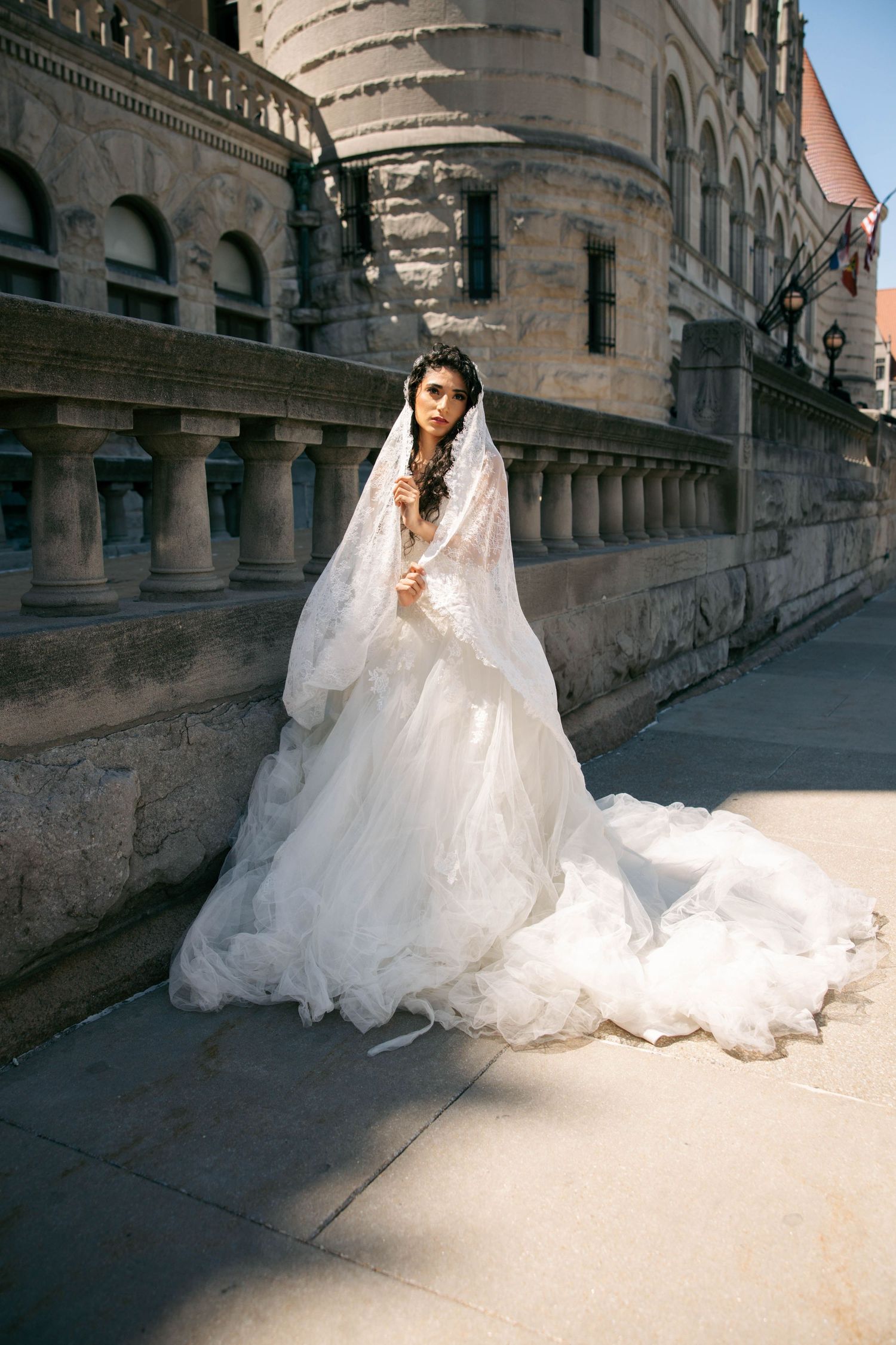 Elegant bride in white lace wedding gown with long veil stands beside historic stone castle.