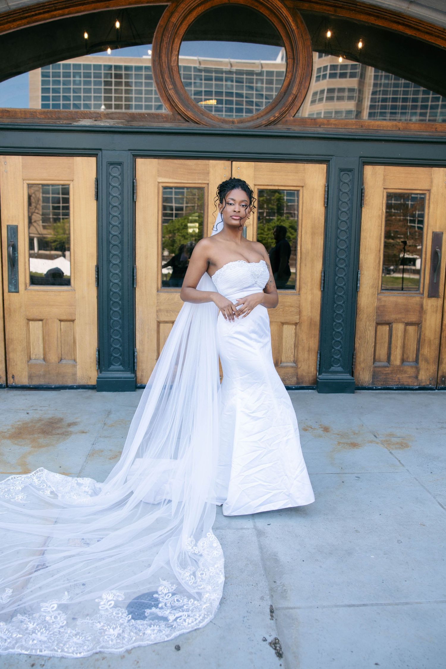 Elegant bride in strapless white gown with lace cathedral train poses before wooden doors.
