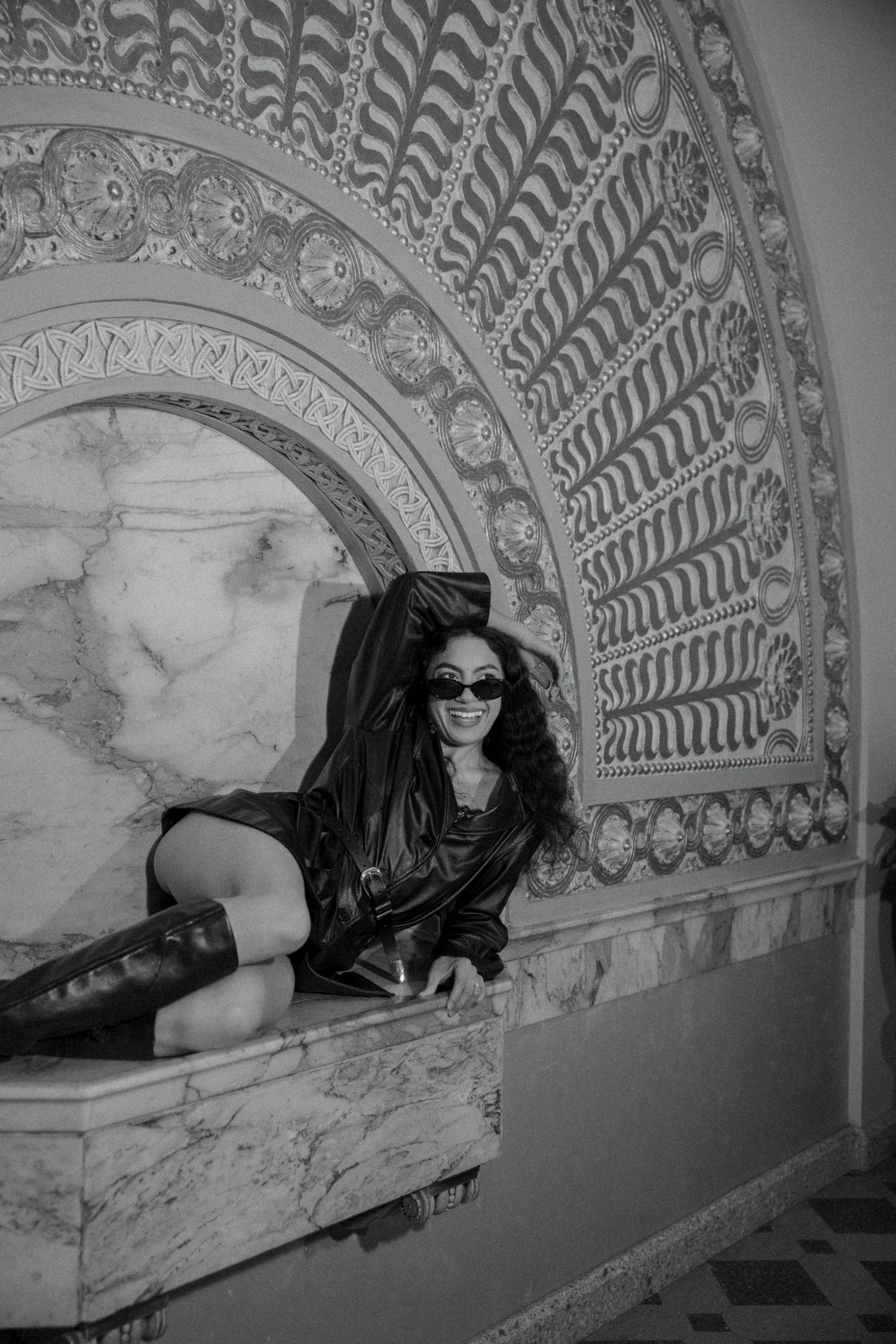 Woman in leather jacket and boots poses in ornate marble archway with intricate mosaic patterns, black and white photo.