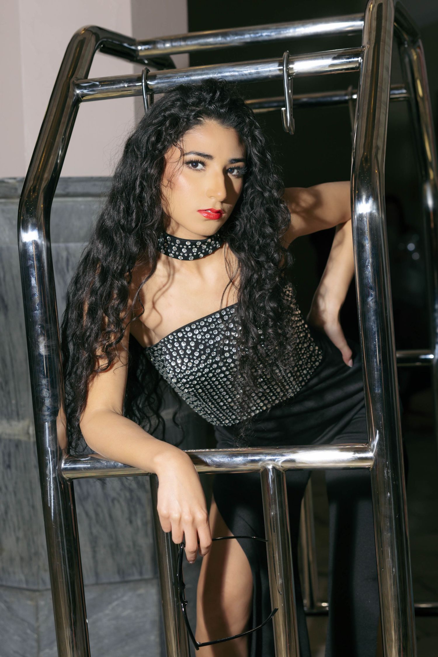 Woman in black studded corset dress with choker posing on chrome luggage cart.