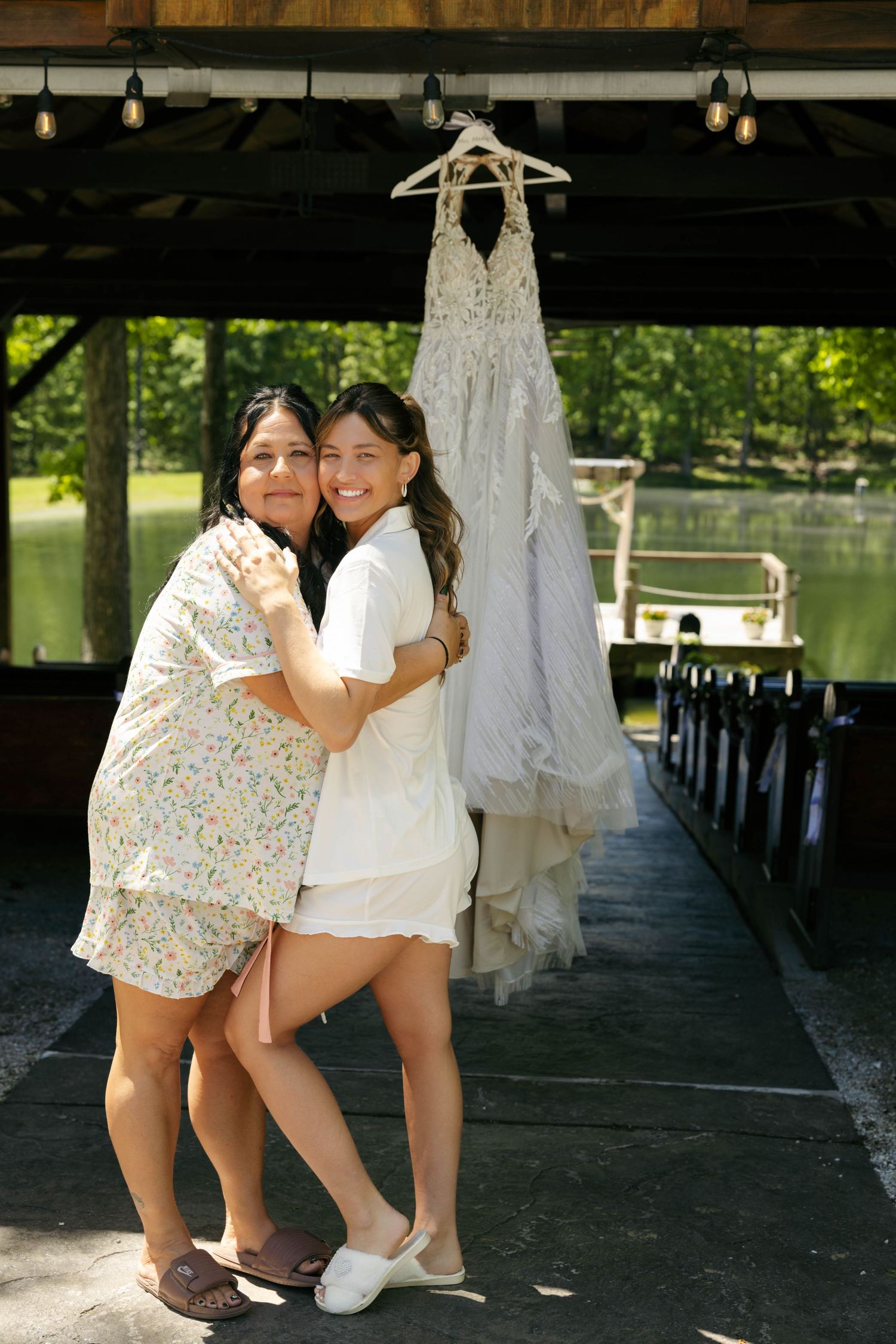 Two women hugging on a dock with a white wedding dress hanging behind them near a serene lake.