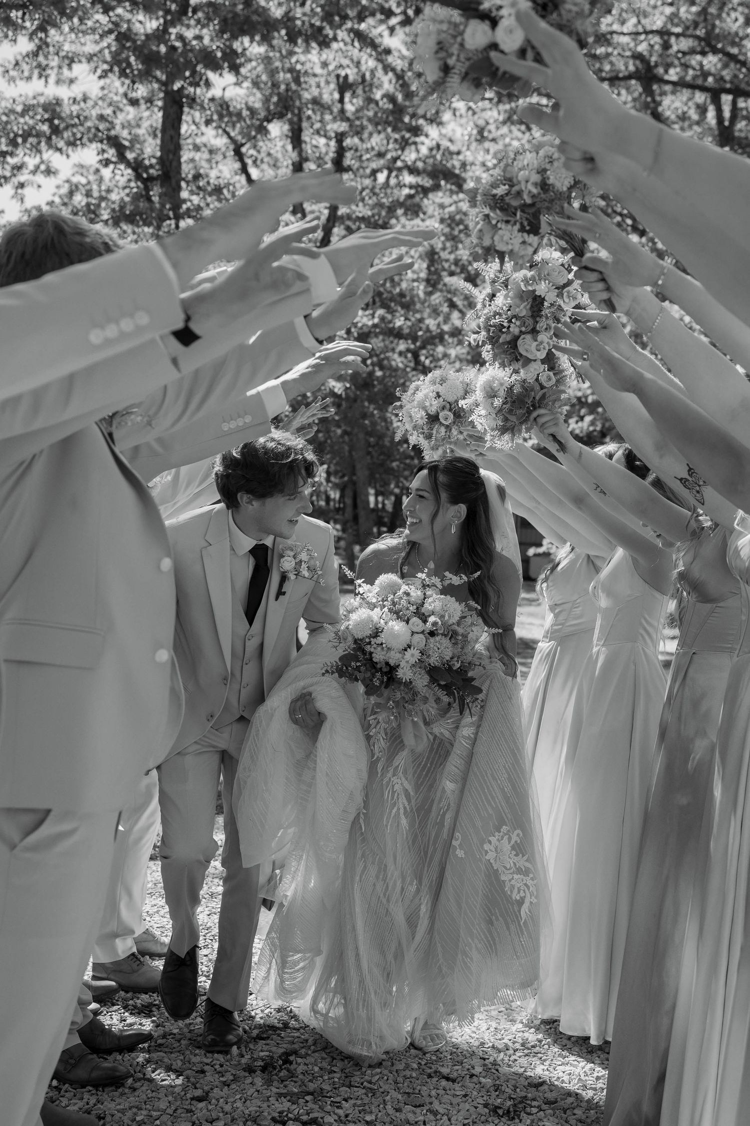 Black and white photo of bride and groom kissing under an arch of hands formed by wedding party outdoors.