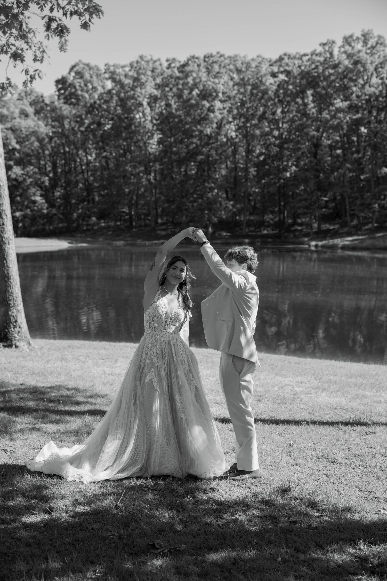 Black and white photo of a bride and groom dancing by a lake surrounded by trees on their wedding day.