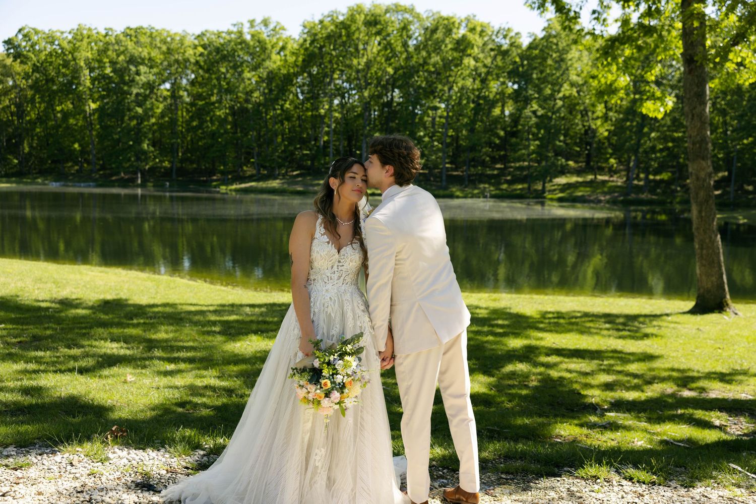 Bride and groom share a kiss near a serene pond surrounded by lush green trees on their wedding day.