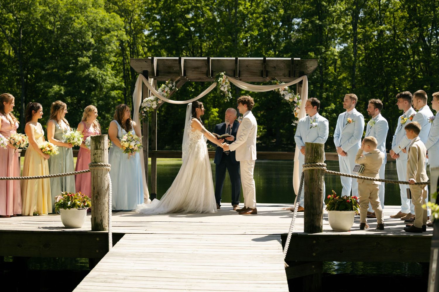 Wedding ceremony on lakeside dock with bridesmaids in pastel dresses and groomsmen under rustic floral arch.