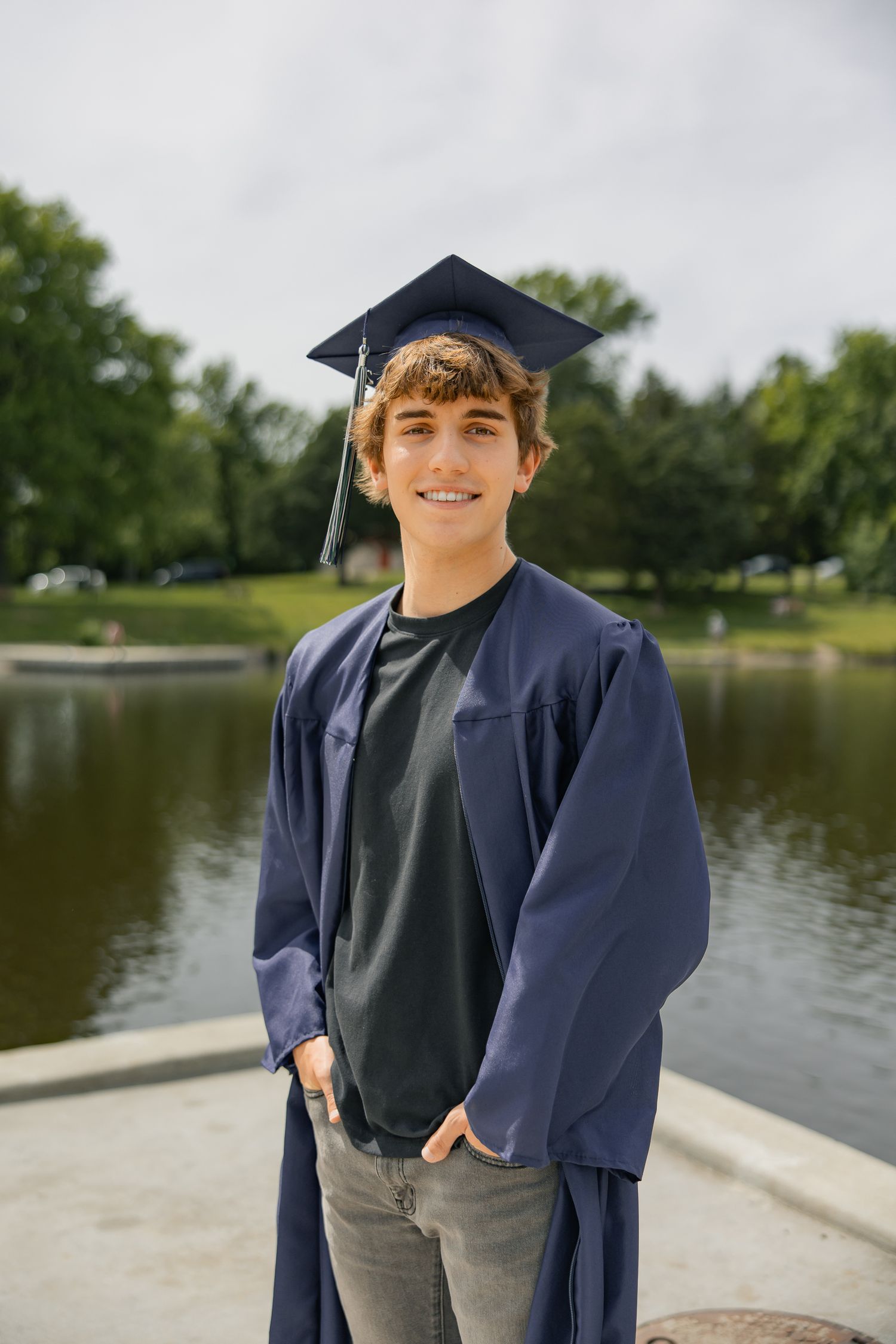 A graduate in navy blue cap and gown smiles by a lakeside setting.