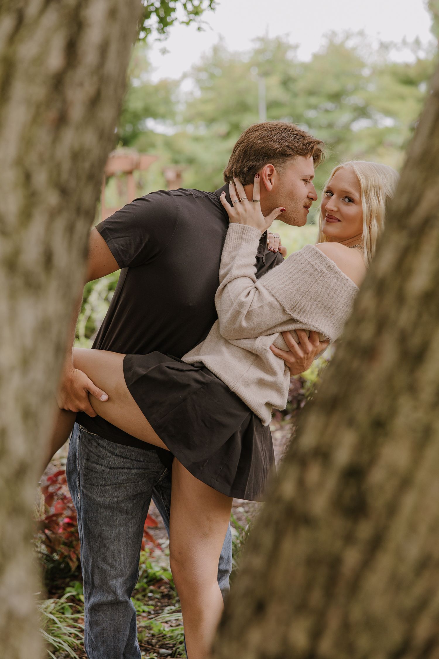 Playful romantic moment captured between trees in an outdoor setting.