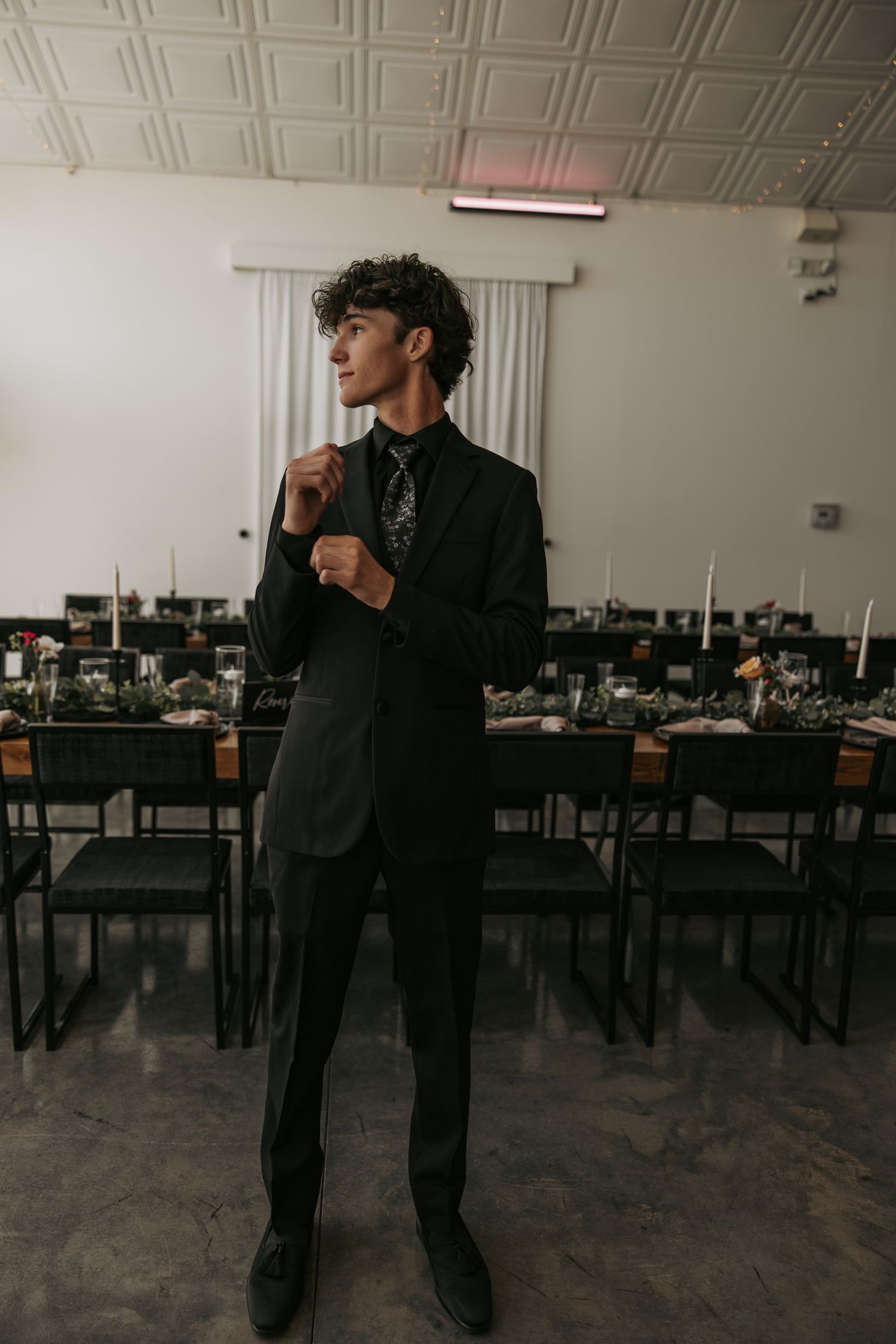 A stylish groom in a black suit stands in a reception hall with empty tables and chairs.