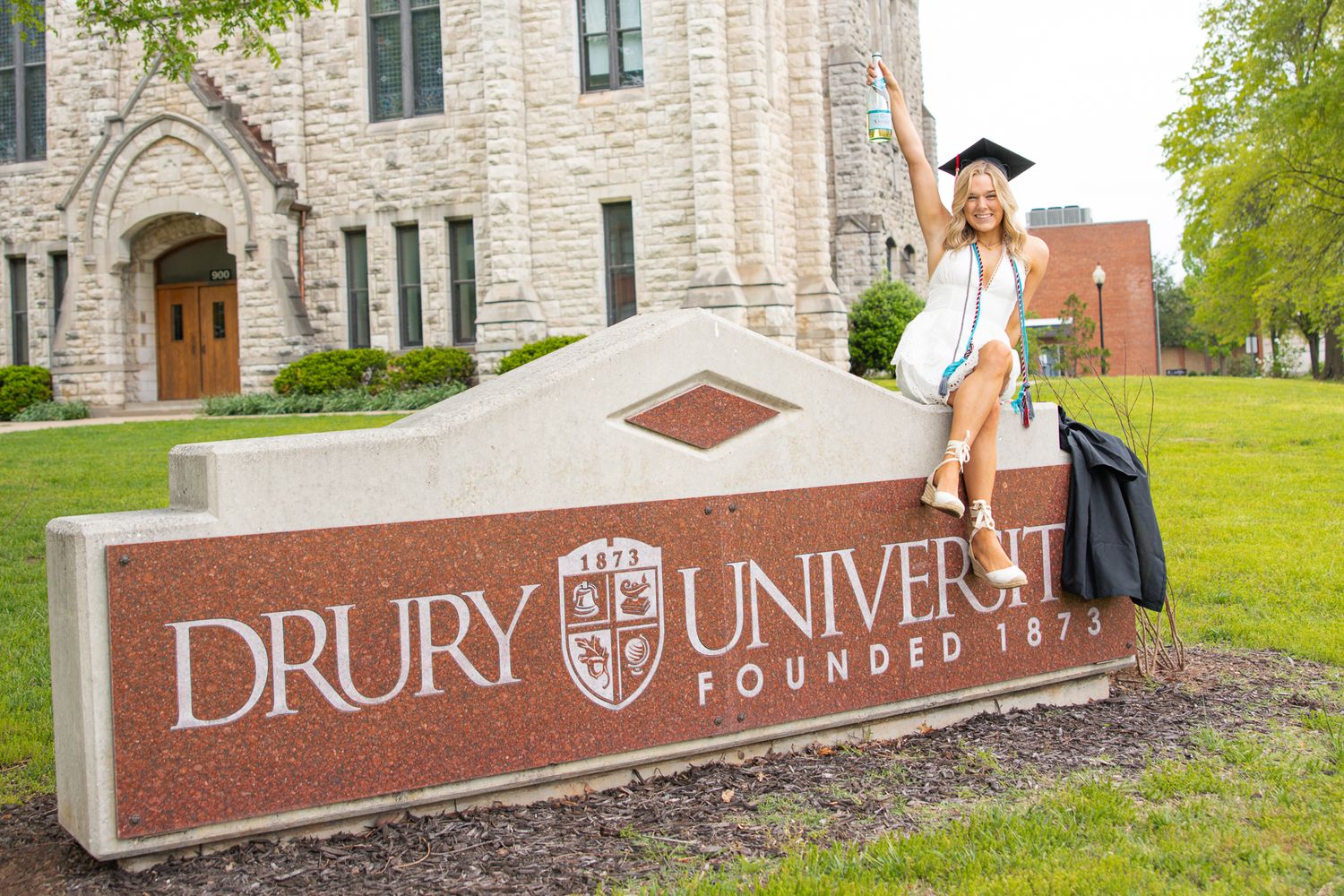 Graduate celebrating by Drury University entrance sign in cap and gown.