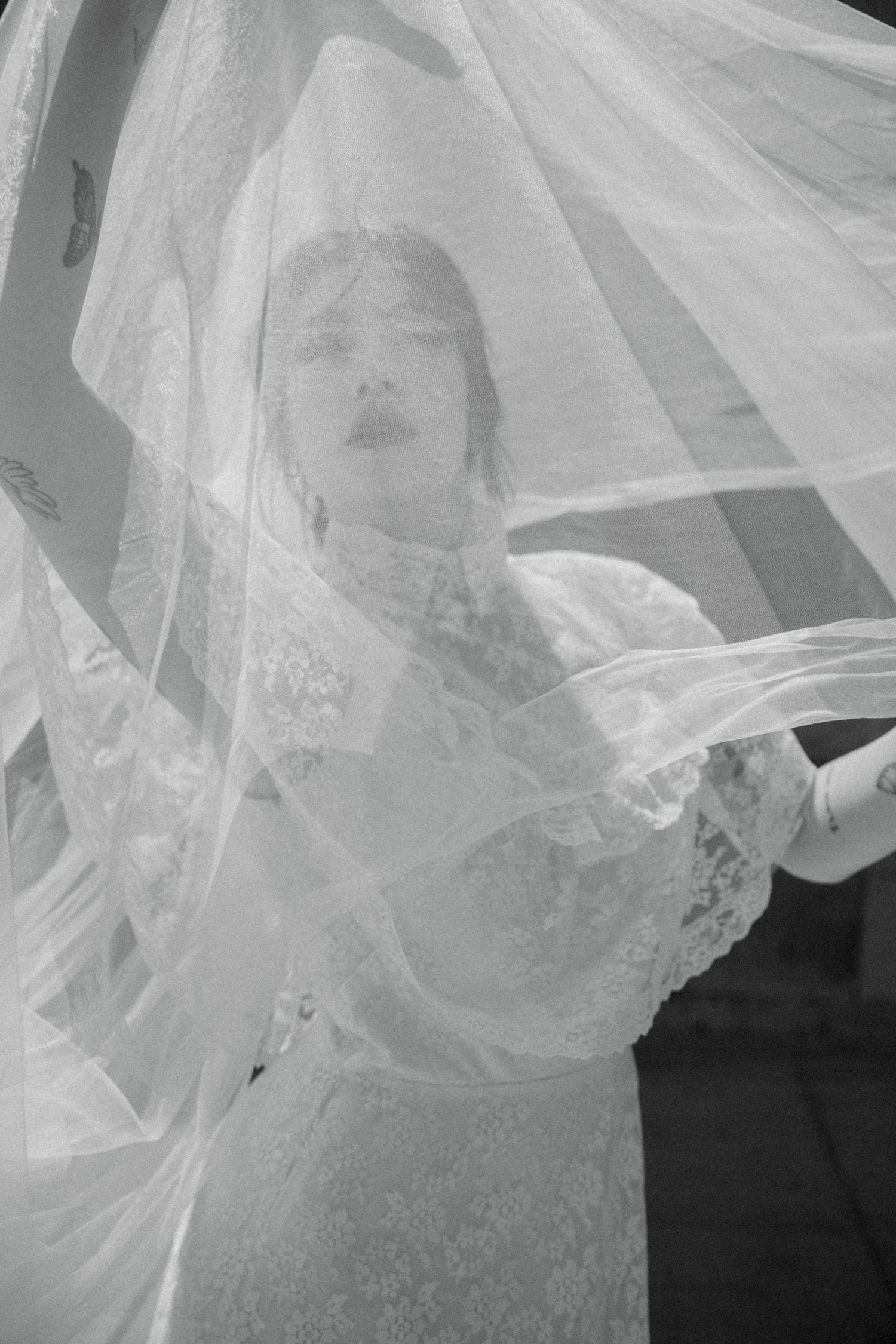 Black and white photo of a bride in lace dress with flowing veil billowing around her face.