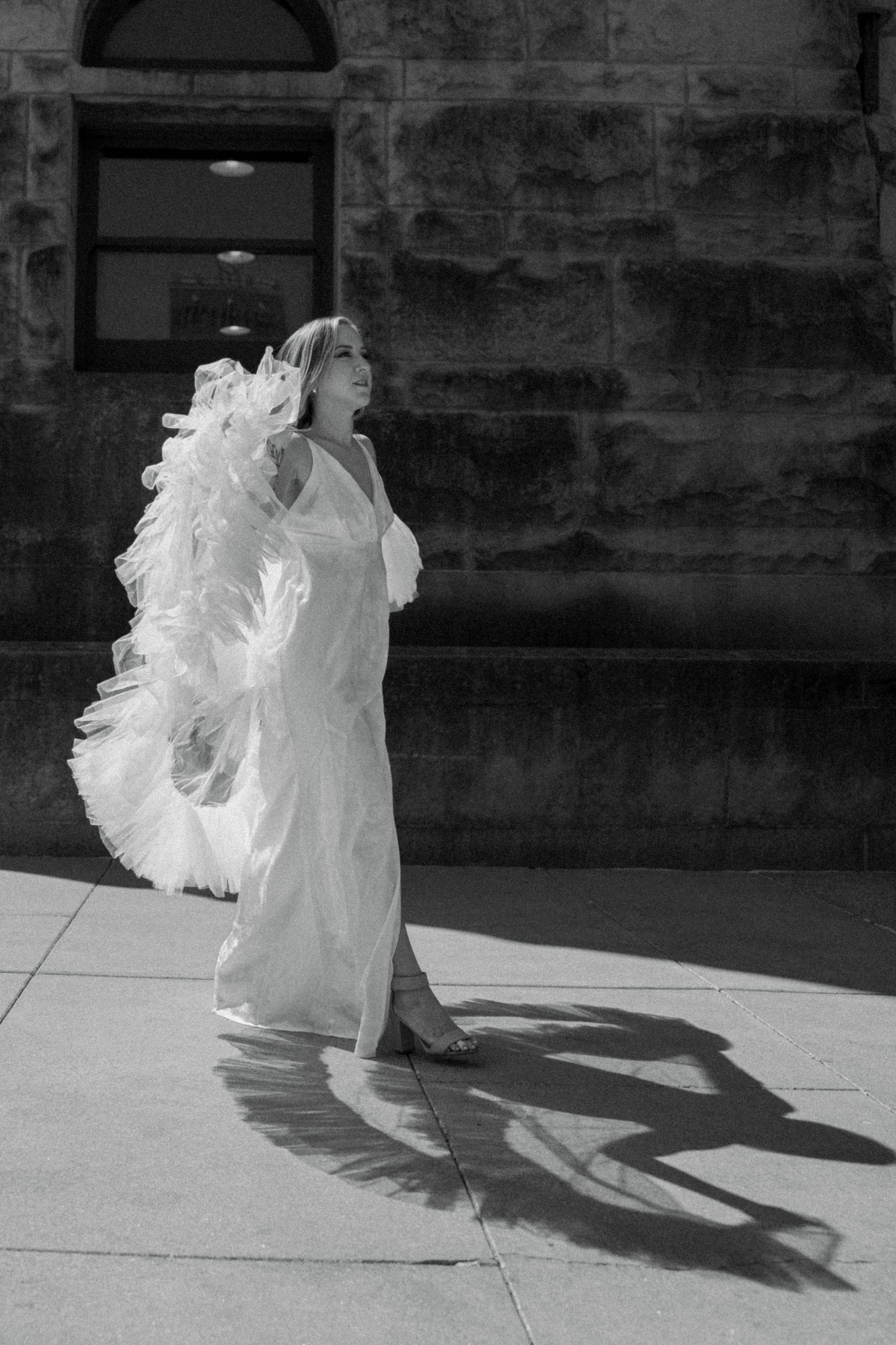 Black and white photo of bride in white dress with dramatic ruffled wrap casting shadow on sunny sidewalk.