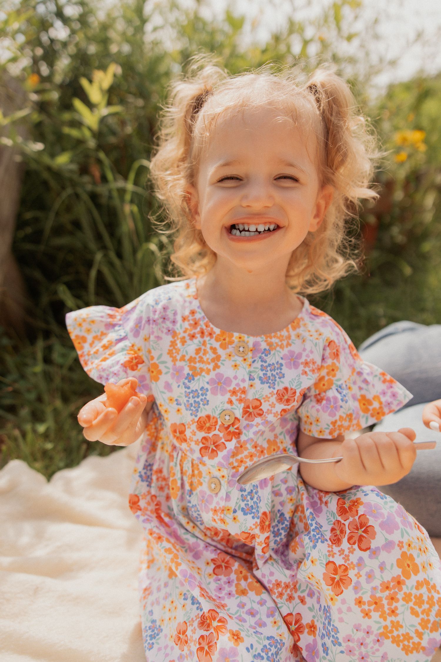 A joyful child in a floral dress plays on a white blanket outdoors on a sunny day.