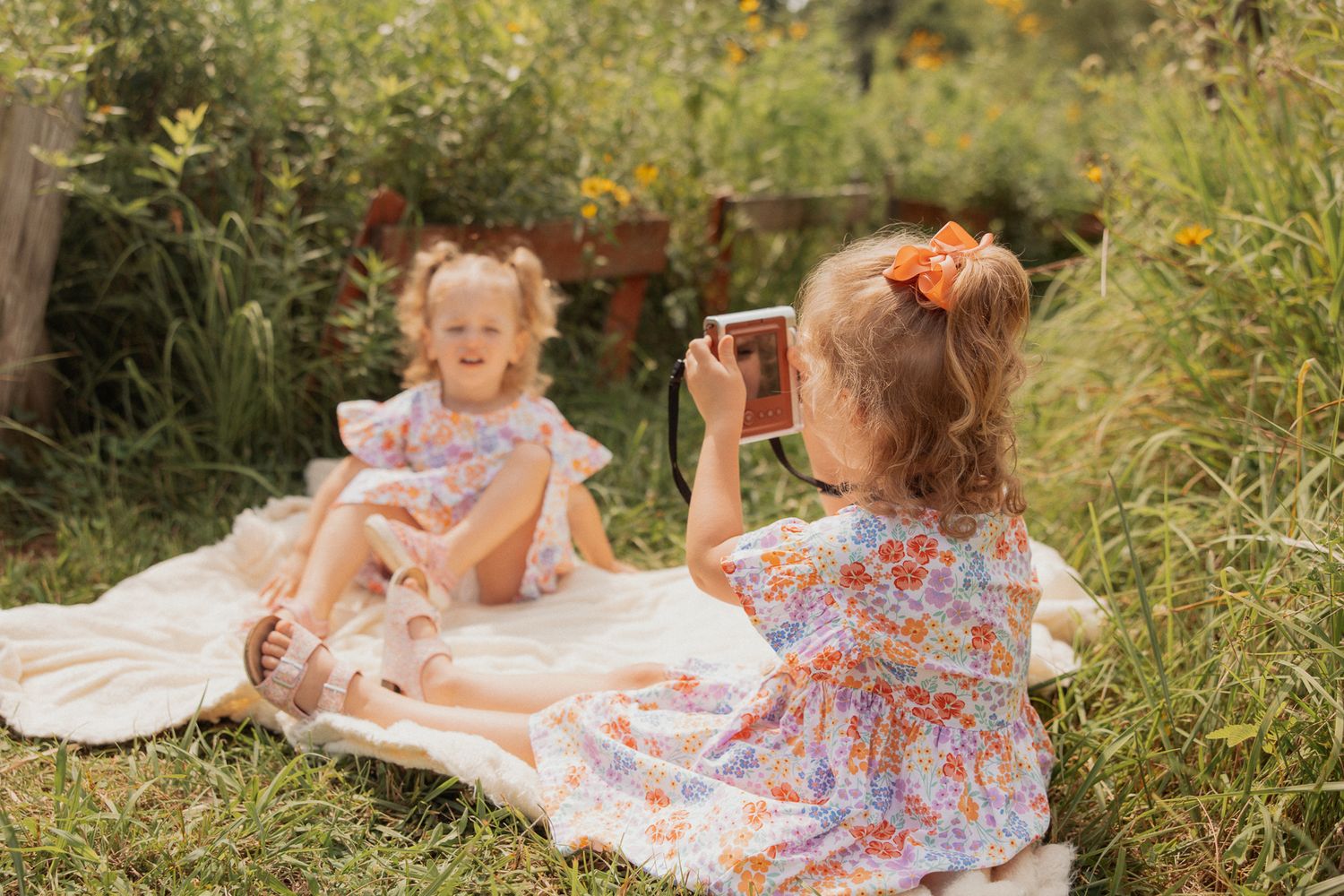 Two in matching floral dresses having a picnic and taking photos on a blanket outdoors.