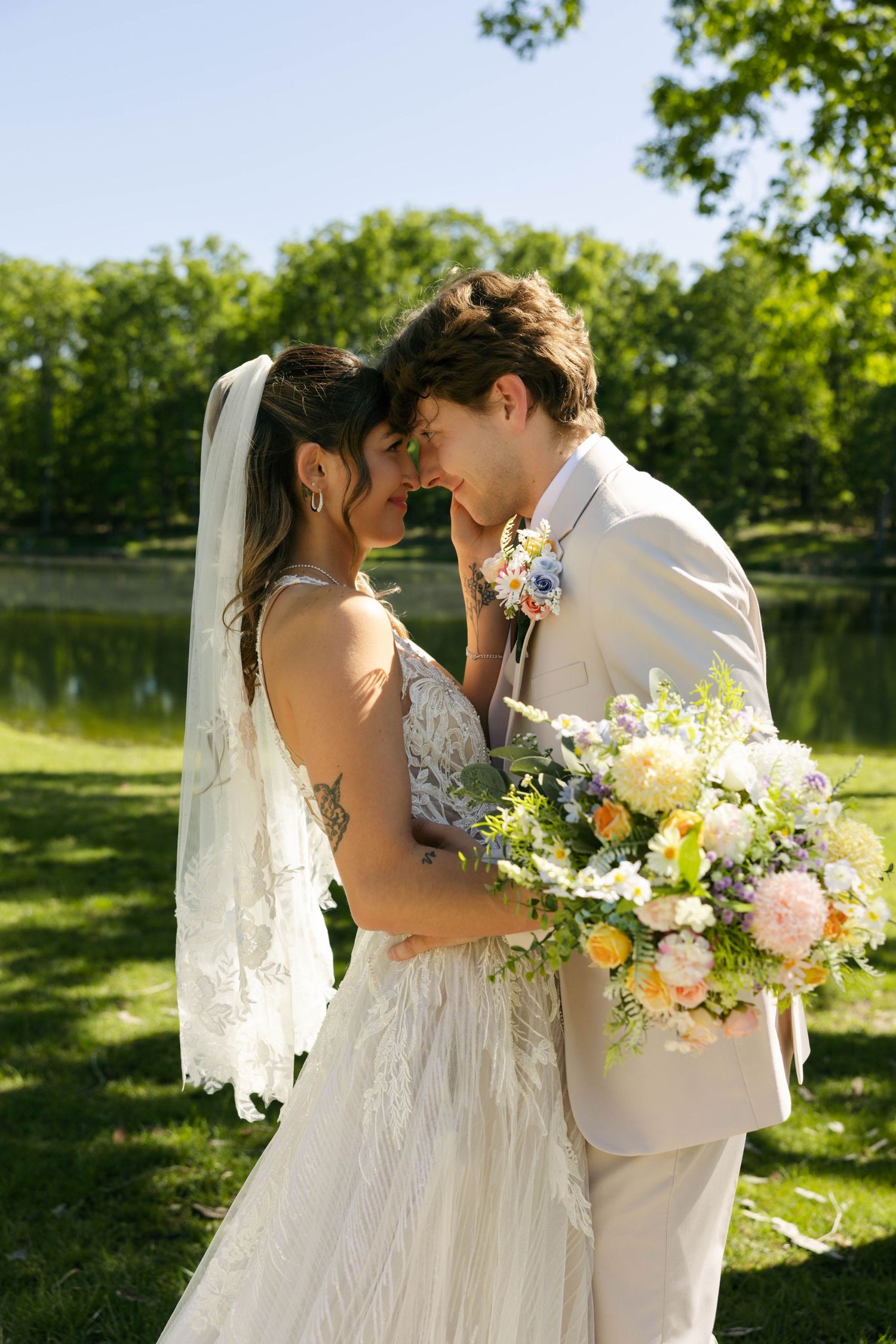 A bride and groom share a romantic forehead touch outdoors, she holds a pastel bouquet, wearing a lace gown and veil.