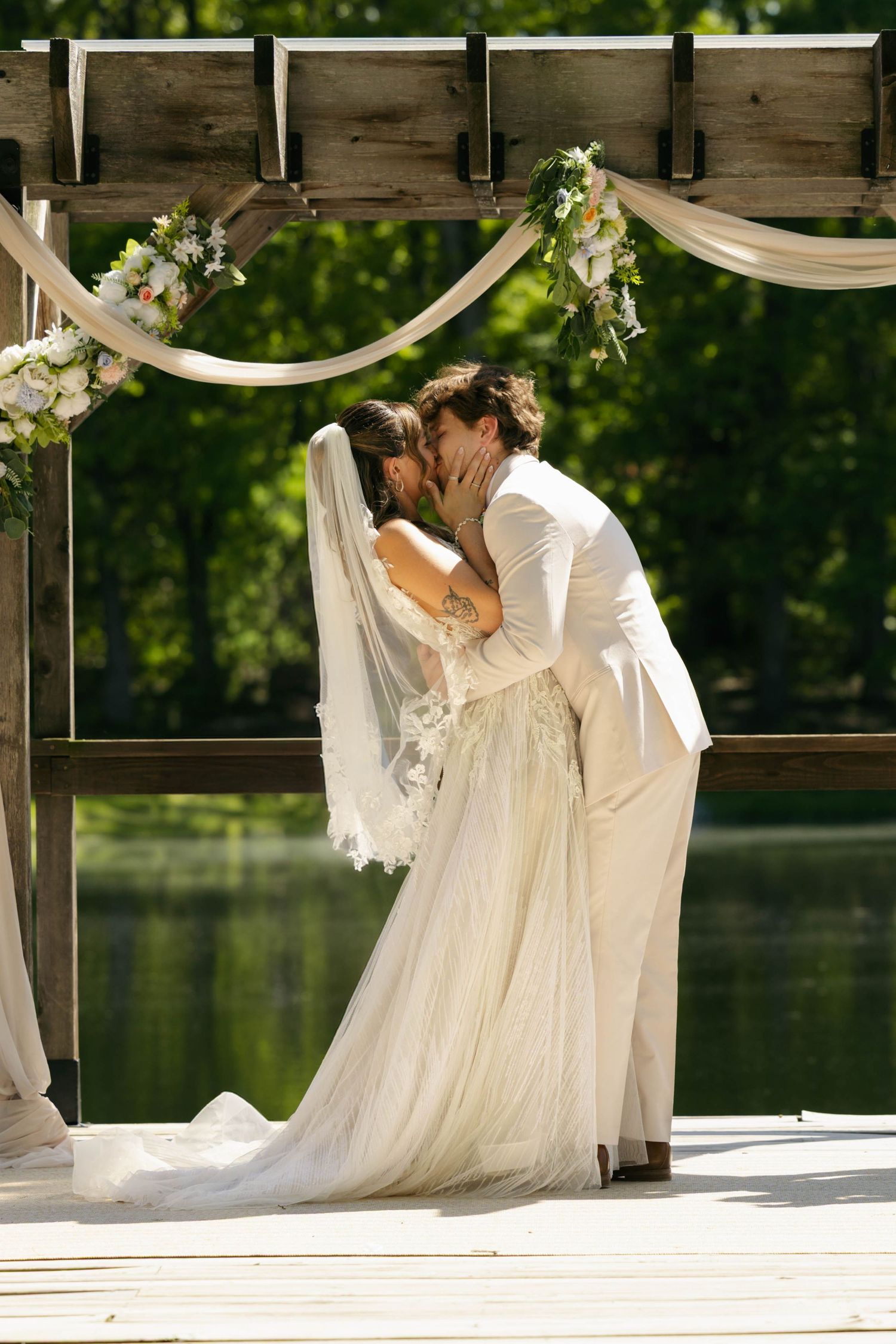 Bride and groom share first kiss under floral arch on waterfront dock during outdoor wedding ceremony.