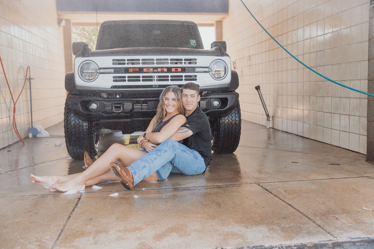 Couple relaxes on concrete floor in front of a customized white Ford Bronco.