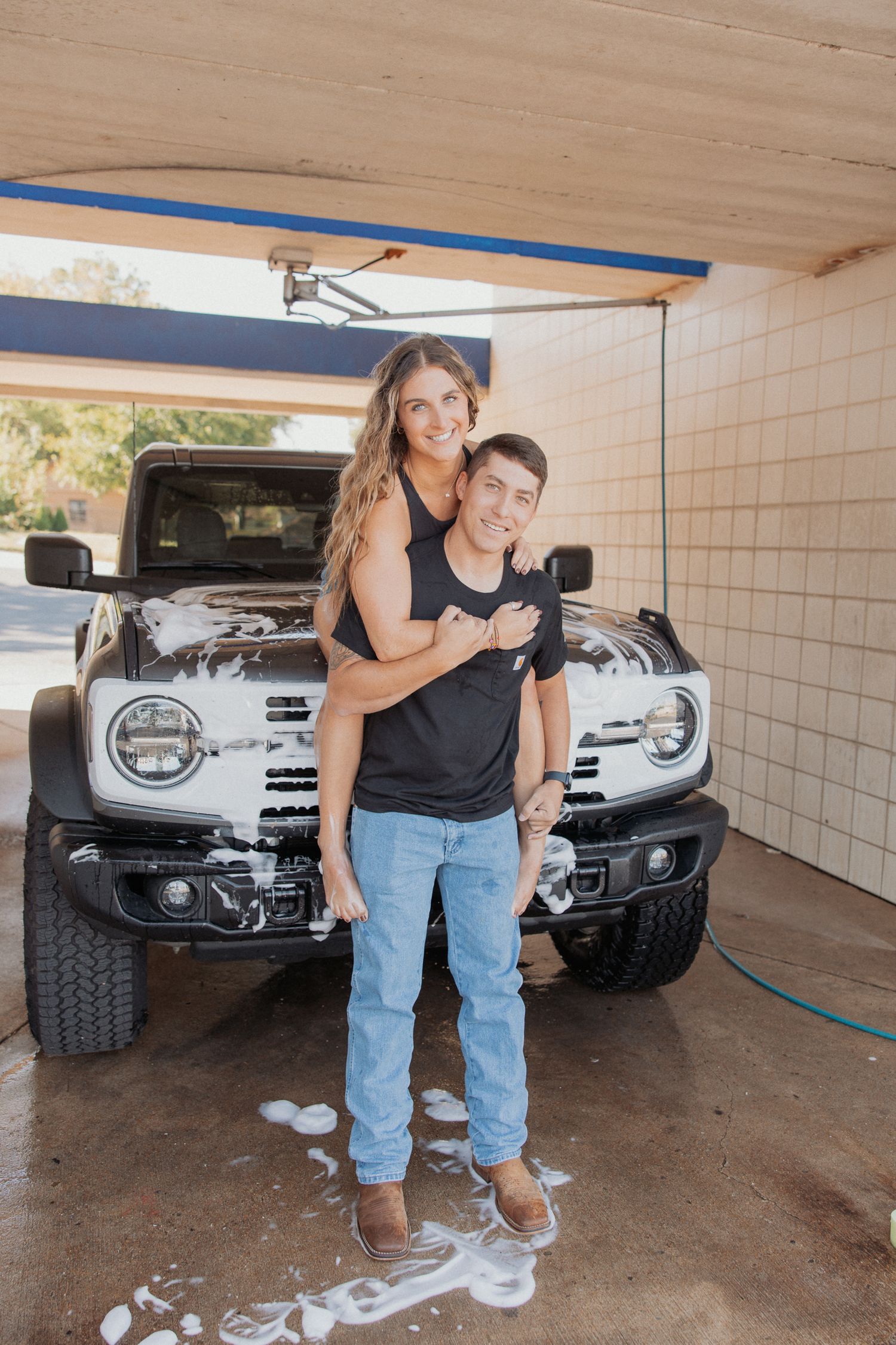 A couple poses playfully in front of a silver truck in a covered parking area.